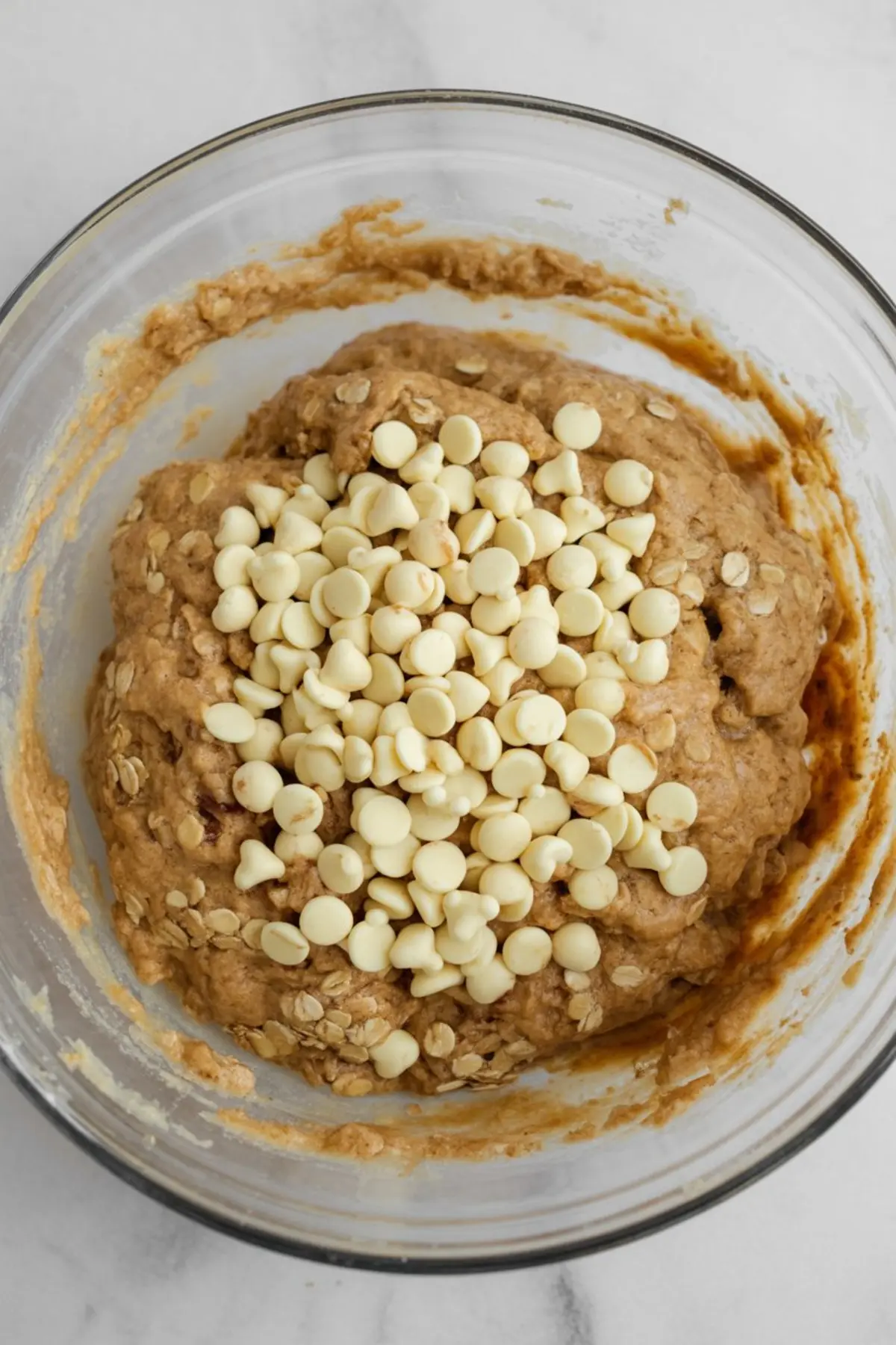 Chunky oatmeal cookie dough mixed with white chocolate chips in a glass bowl, highlighting the thick texture and visible oats.
