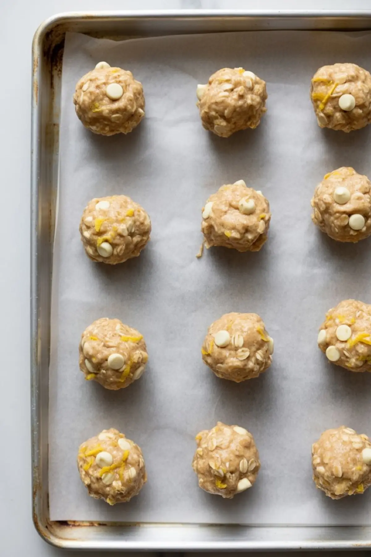Unbaked lemon oatmeal cookie dough balls placed in rows on a parchment-lined baking sheet, each topped with oats, lemon zest, and white chocolate chips.

