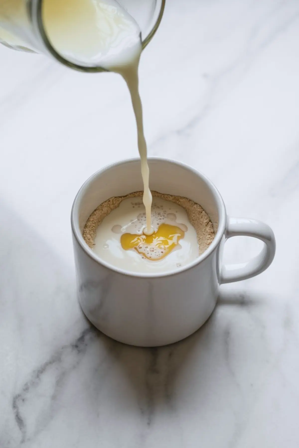 Close-up of milk being poured into a white mug containing lemon protein mug cake dry ingredients, showing liquid pooling over protein powder and sweetener on a marble surface.
