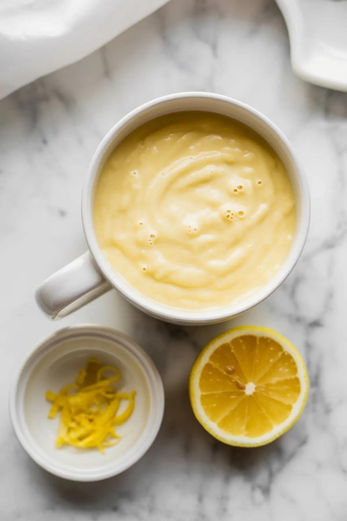 Top-down view of mixed lemon protein mug cake batter inside a white mug, showing smooth pale yellow texture with lemon zest beside a halved lemon on a marble countertop.
