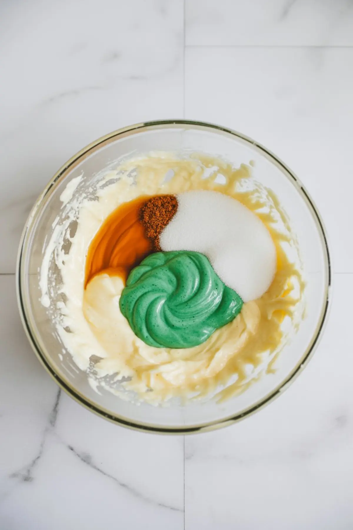 Glass mixing bowl holding whipped butter mixture with sugar, vanilla extract, peppermint flavoring, and green food coloring, placed on a white marble countertop.
