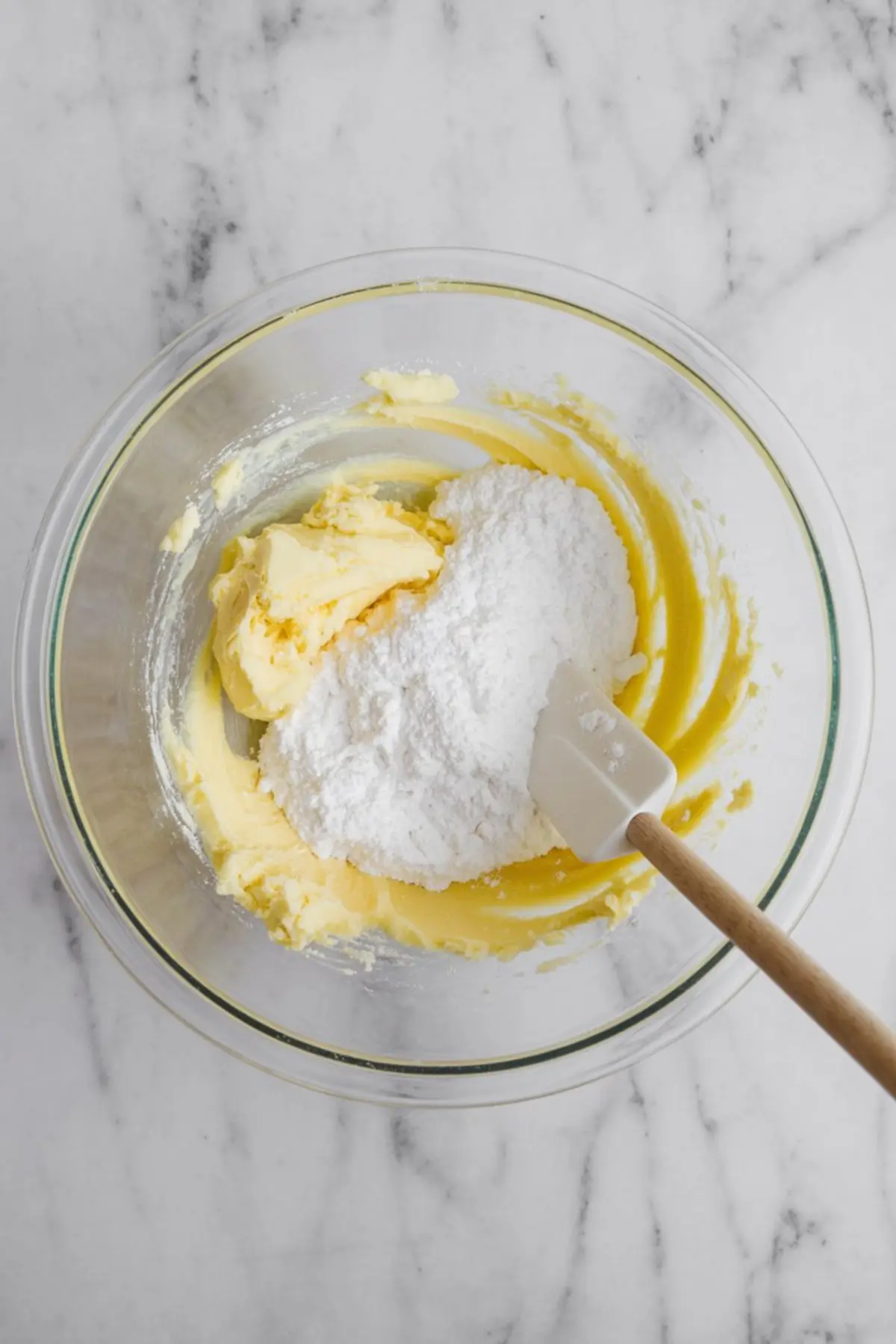 Creamy butter mixture in a clear glass bowl with a spatula, topped with a pile of powdered sugar, prepared on a white marble surface.
