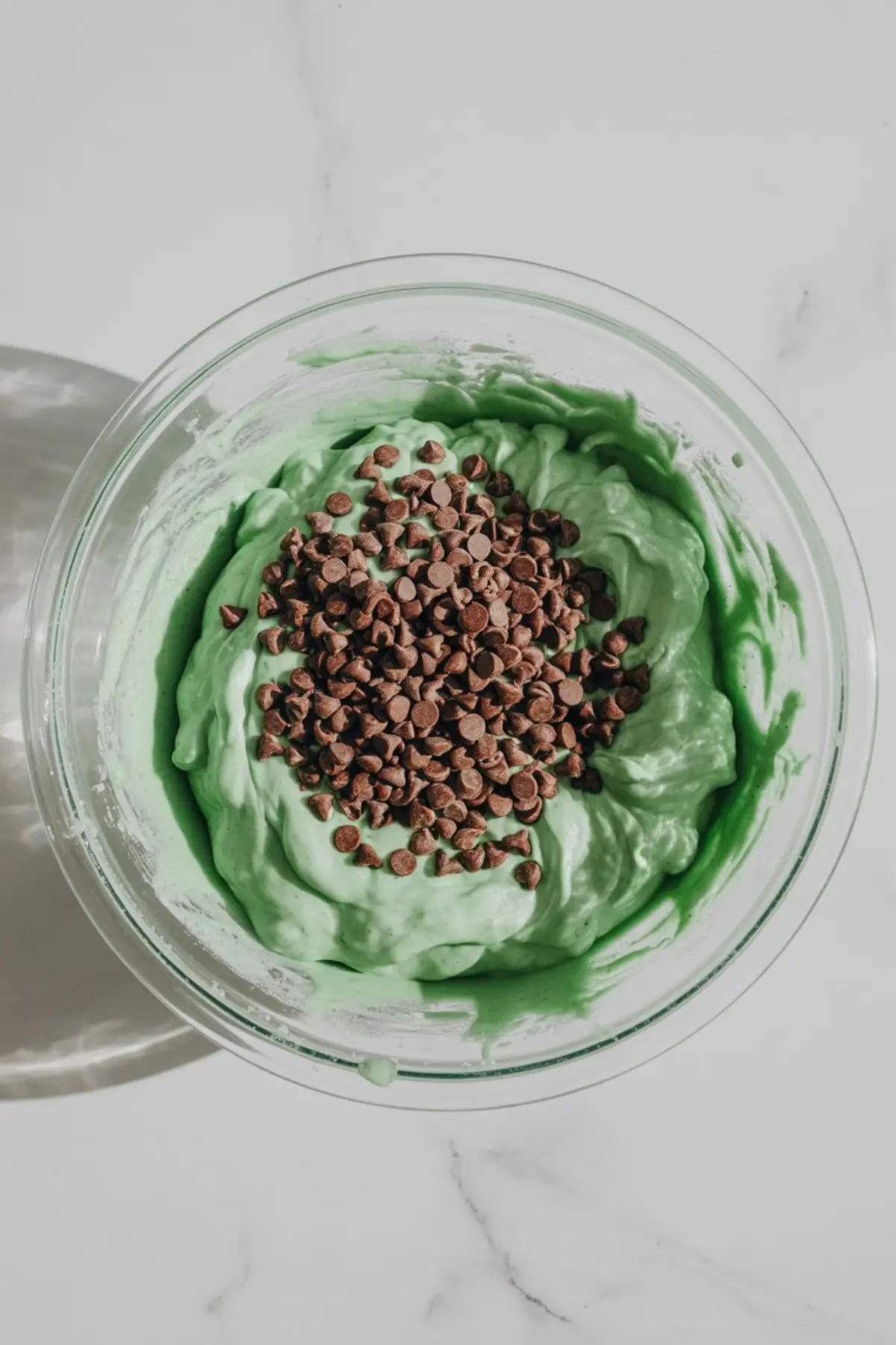 Green-tinted cookie dough blended with mini chocolate chips in a glass mixing bowl, ready for shaping into snowball cookies.

