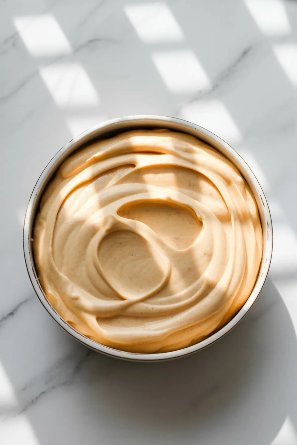 Top-down view of freshly whipped orange cardamom frosting with swirled texture inside a round cake pan on a marble surface.
