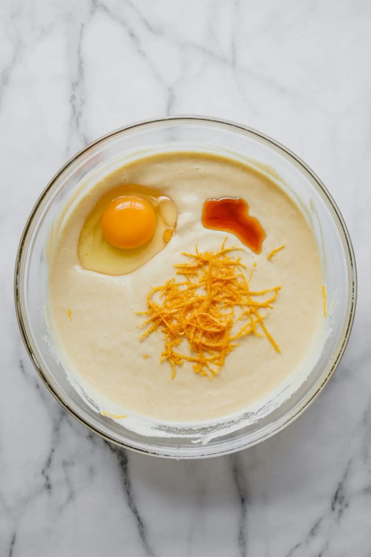 Cake batter in a glass bowl with fresh egg, orange zest, and vanilla extract added on top, ready to be blended for orange cardamom cake.