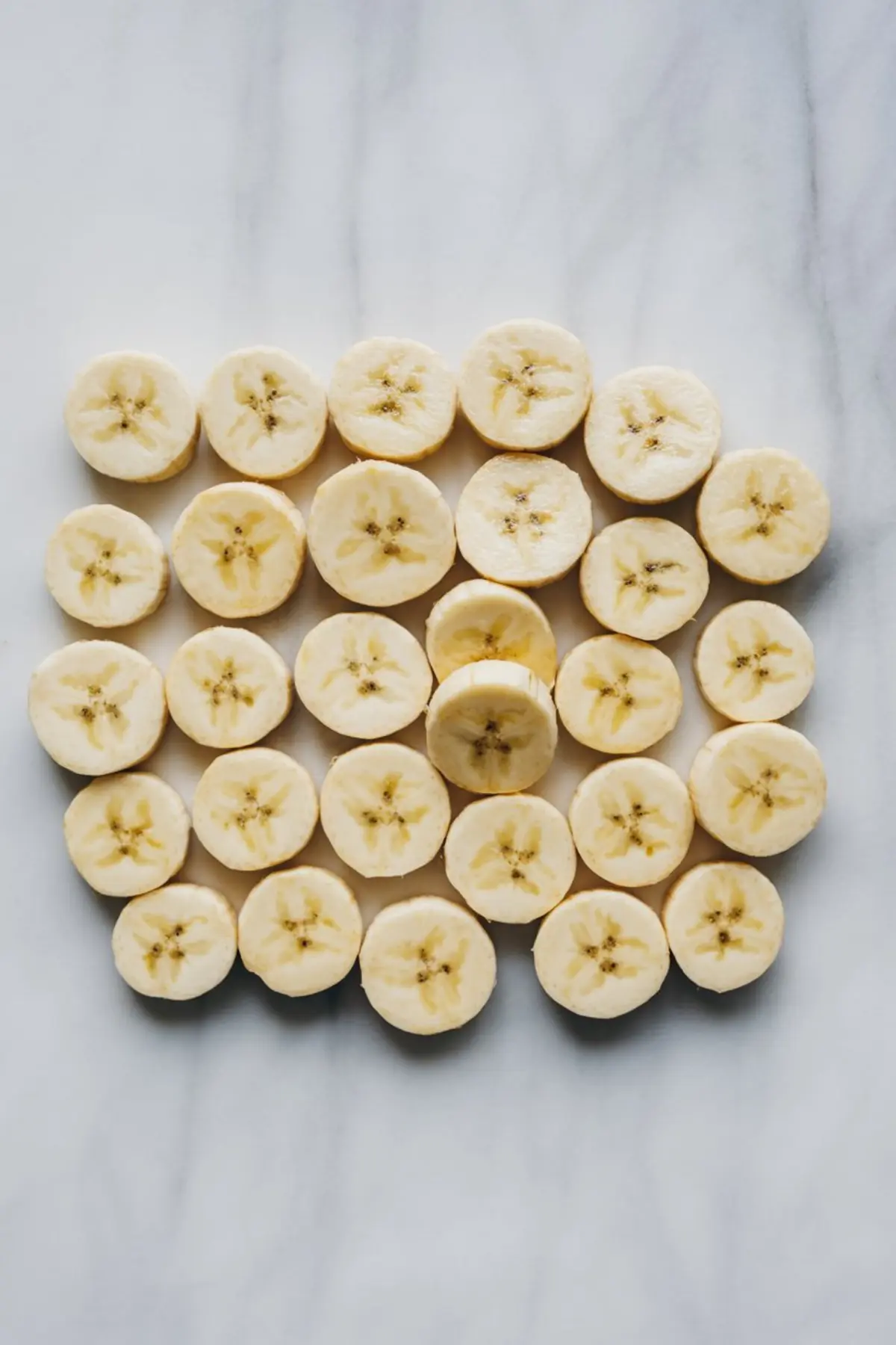 Overhead image of neatly arranged banana slices on a marble countertop, showcasing their fresh texture and uniform cut for snack preparation.

