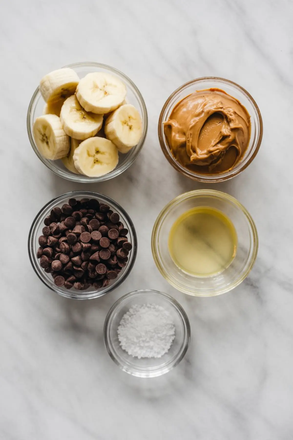 Flat lay of five small glass bowls holding ingredients for banana bites: sliced bananas, creamy peanut butter, dark chocolate chips, coconut oil, and flaky sea salt, all arranged on a marble background.
