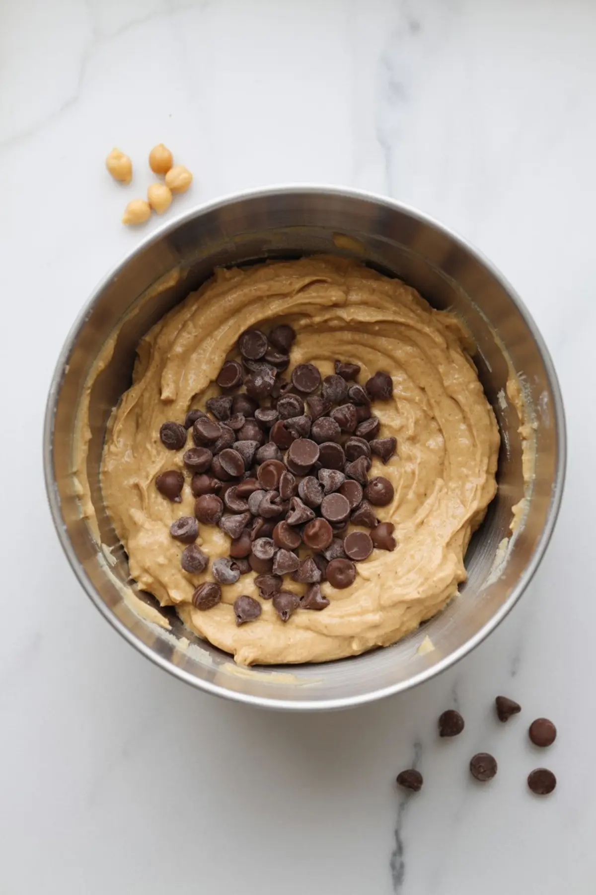 Metal mixing bowl with peanut butter cookie dough topped with semi-sweet chocolate chips on a white marble surface, with a few chickpeas and chocolate chips scattered beside it.
