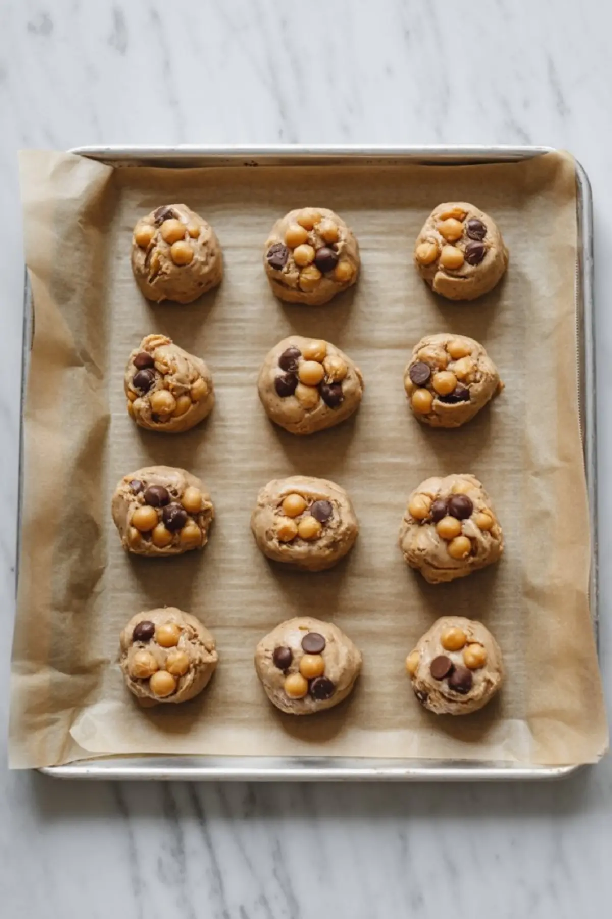 Raw cookie dough scoops with chickpeas and chocolate chips arranged on a parchment-lined baking tray, ready to bake.
