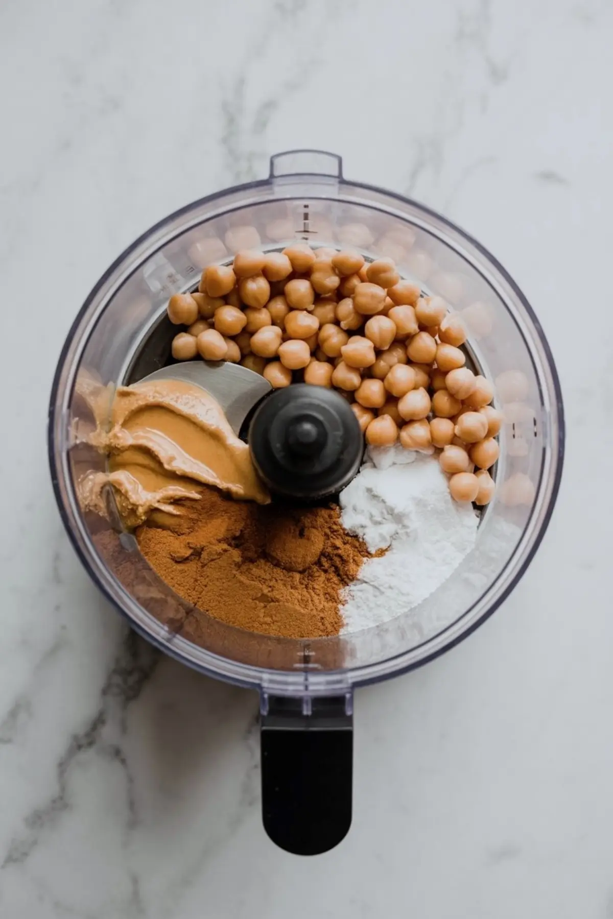 Overhead view of a food processor filled with chickpeas, peanut butter, cinnamon, and baking soda, placed on a marble surface.
