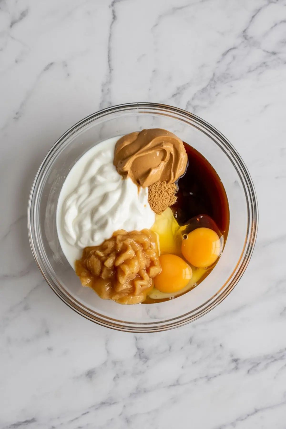Glass bowl filled with mashed banana, Greek yogurt, eggs, peanut butter, maple syrup, and coconut sugar on a marble countertop for a protein brownie recipe.