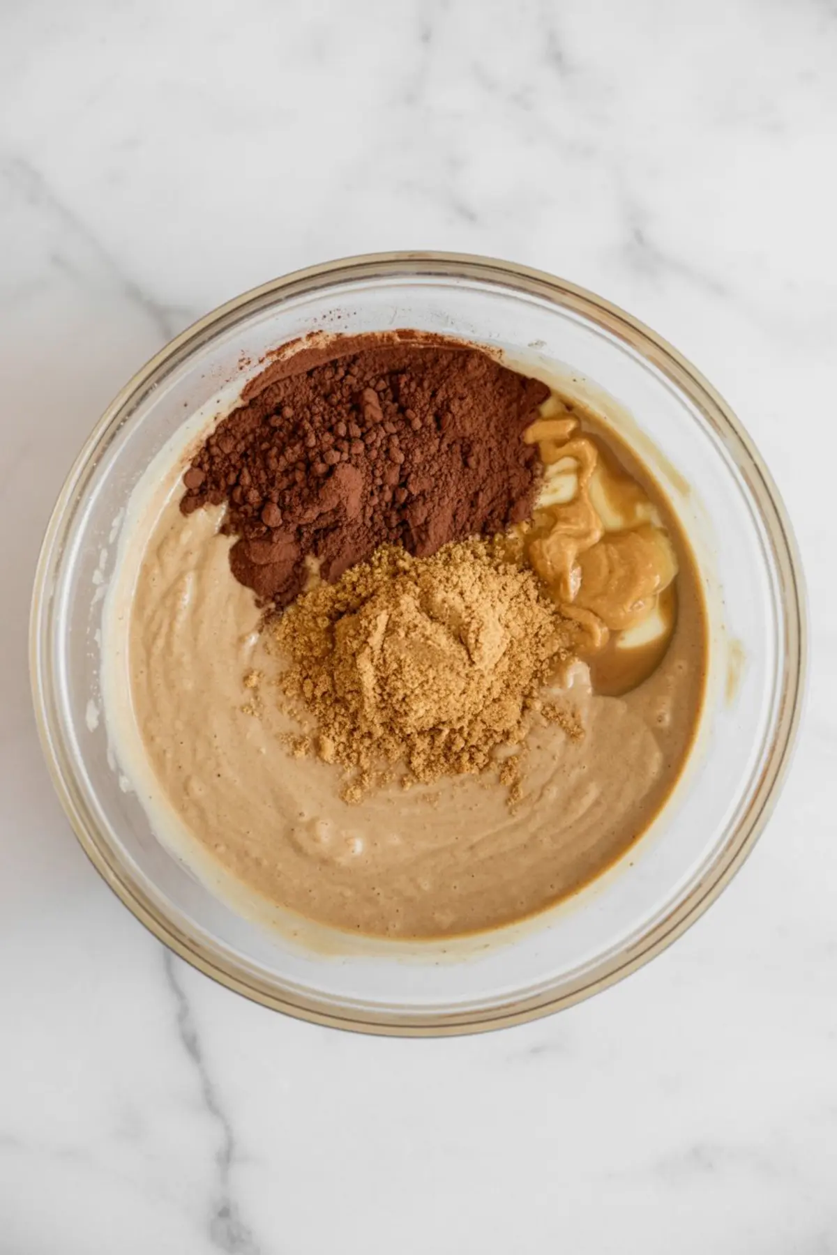 Batter mixture in a glass bowl with cocoa powder, peanut butter, and coconut sugar sitting on top, ready to be blended into a chocolate protein brownie mix.