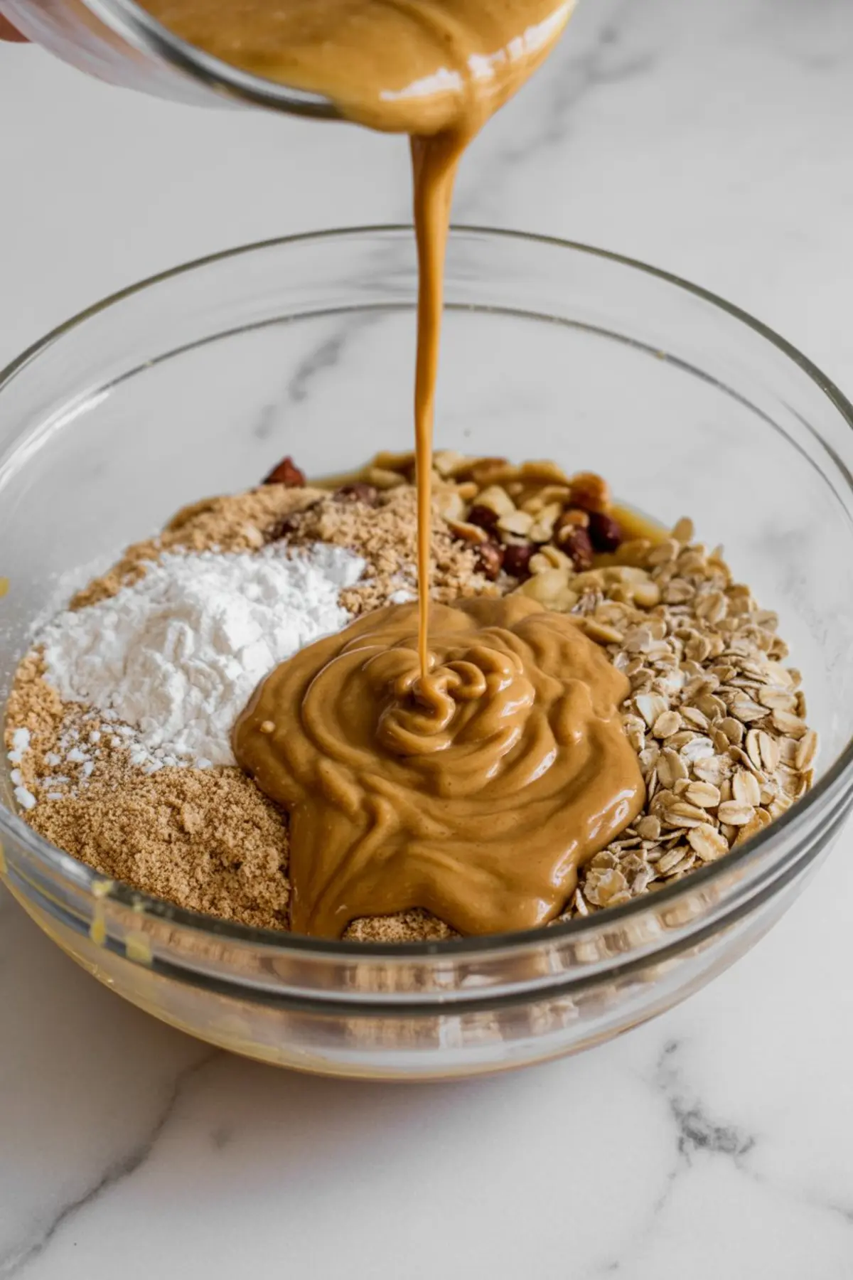 Creamy peanut butter being poured into a glass bowl filled with oats, protein powder, crushed nuts, and powdered sweetener for healthy snack prep.