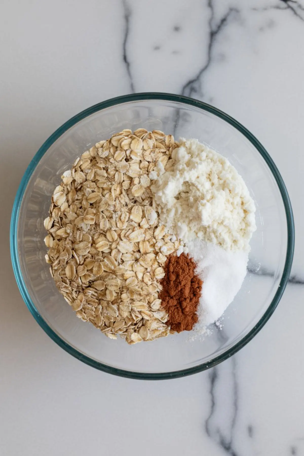 Top-down view of a glass bowl containing rolled oats, coconut flour, cinnamon, and salt, set on a white marble background for protein oat mini mix.