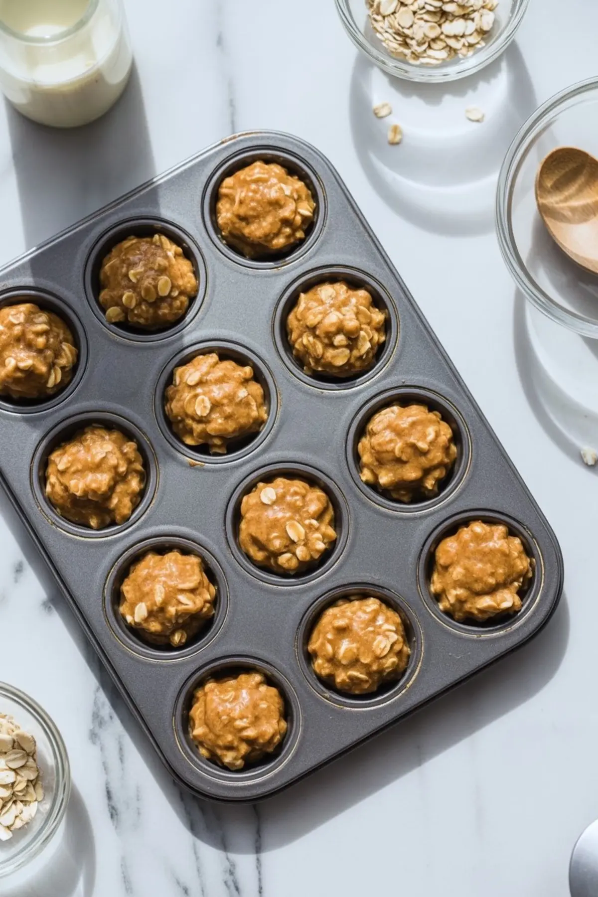 Peanut butter oat mixture scooped into a non-stick mini muffin pan, ready for chilling, with bowls of oats and milk nearby on a marble counter.