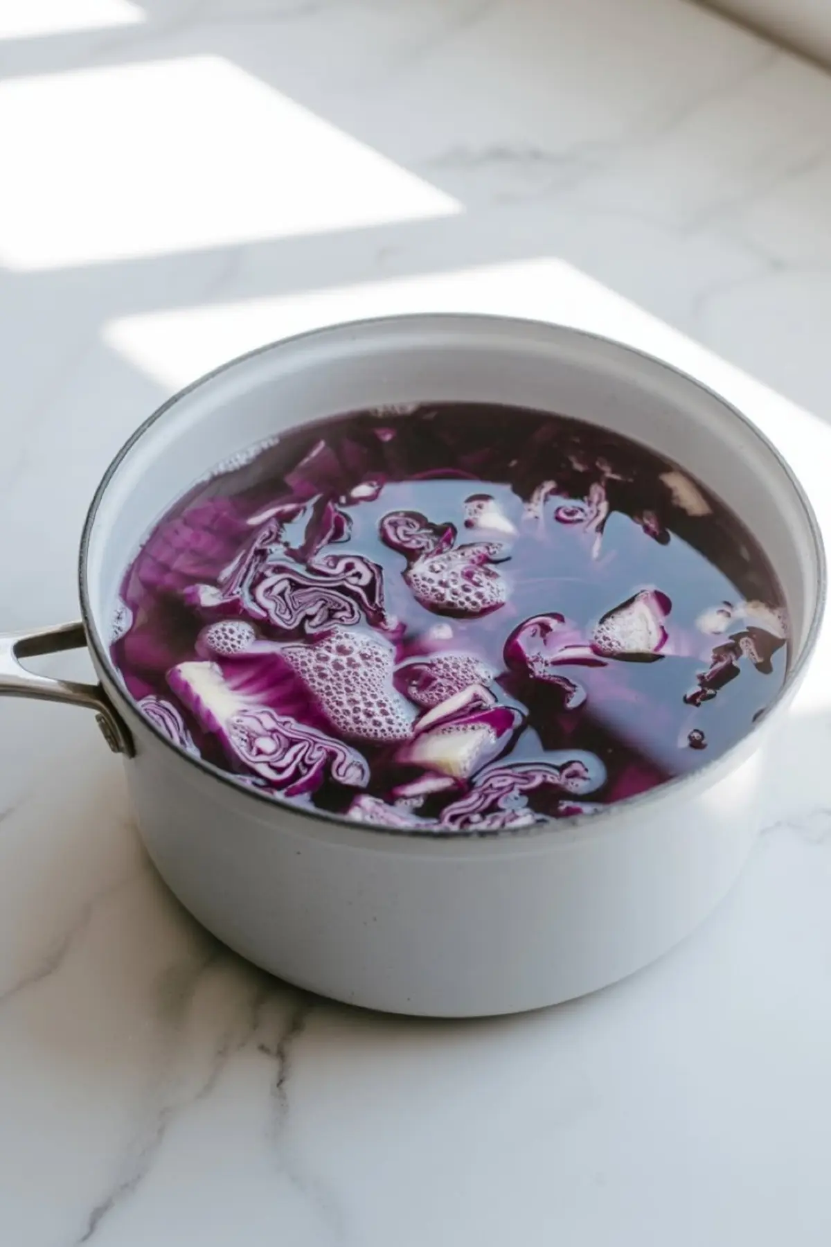 Chopped red cabbage boiling in water inside a white saucepan, steeping to extract natural purple coloring for the panna cotta preparation.
