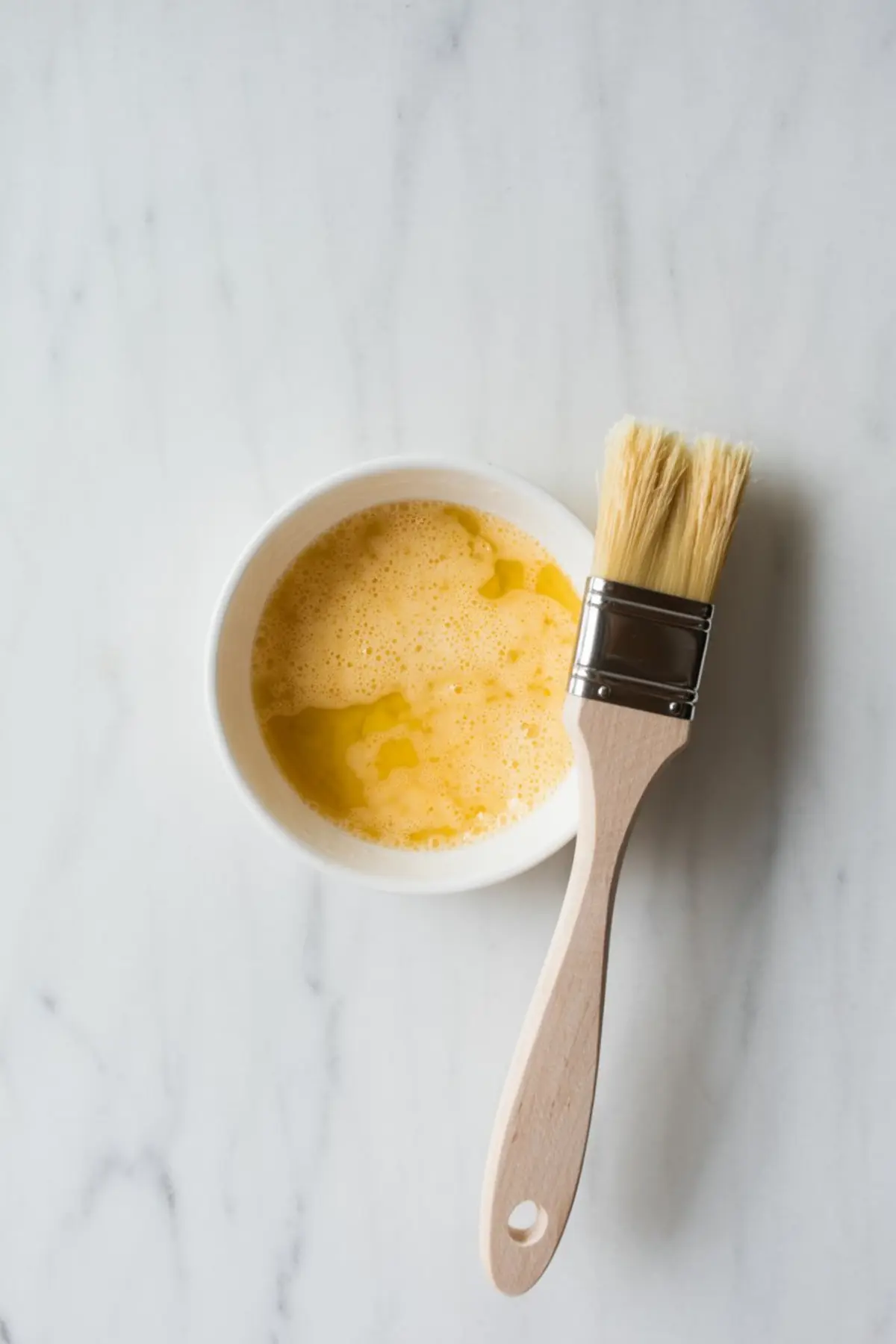 A white ceramic bowl filled with whisked egg wash sits on a marble surface beside a wooden pastry brush with natural bristles.