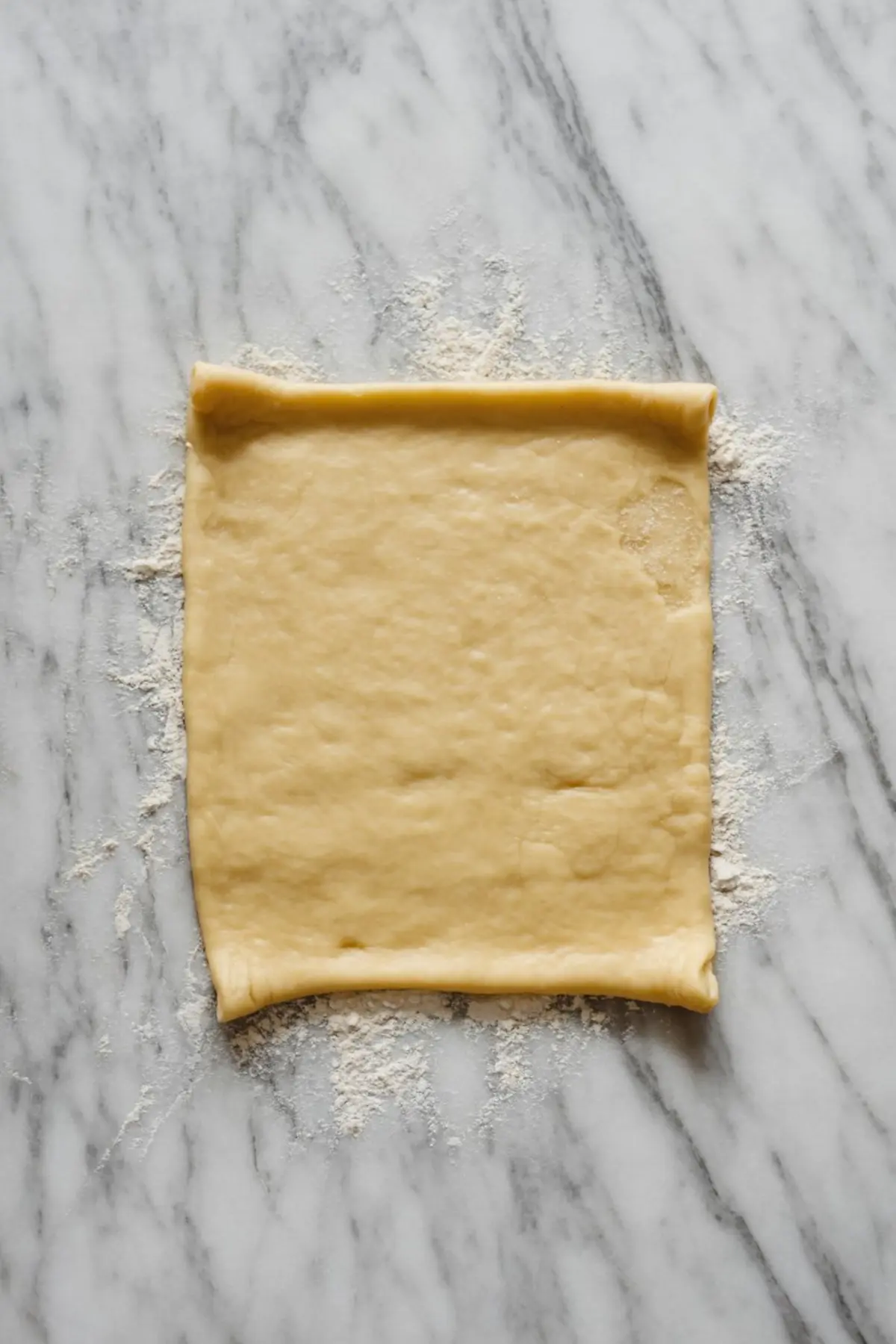 A square sheet of rolled out puff pastry dough is lightly floured on a gray marble countertop, prepared for shaping or filling.