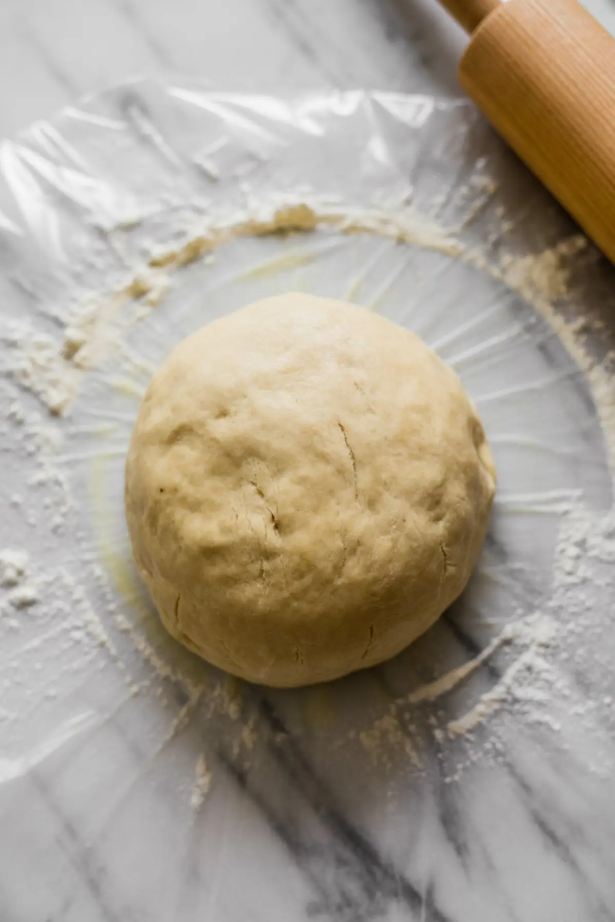 Ball of homemade pie dough on floured parchment, placed on a marble surface next to a wooden rolling pin, ready for rolling and shaping for baking.
