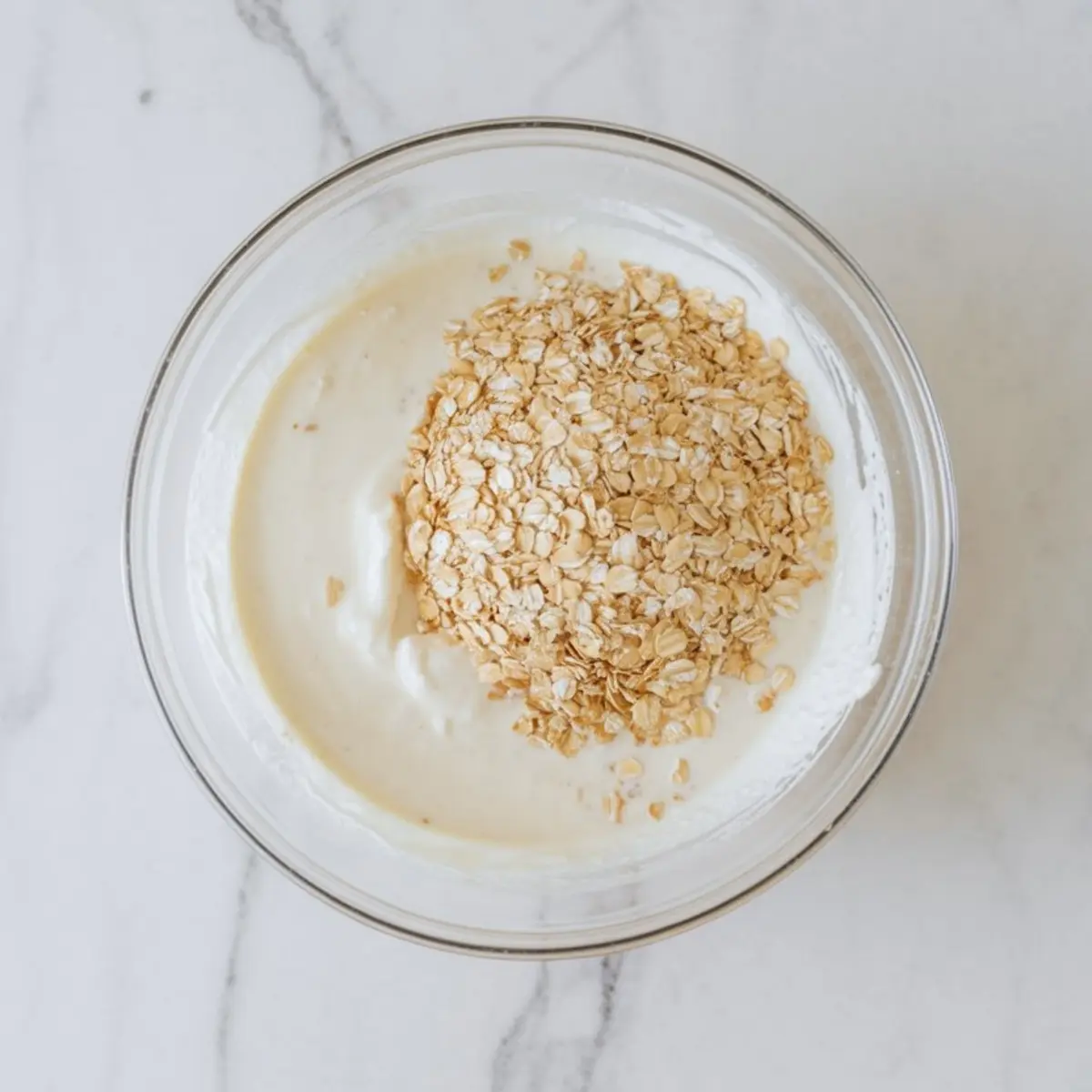 Glass mixing bowl filled with creamy yogurt and a generous portion of rolled oats on a white marble countertop, showcasing the base preparation for overnight oats.