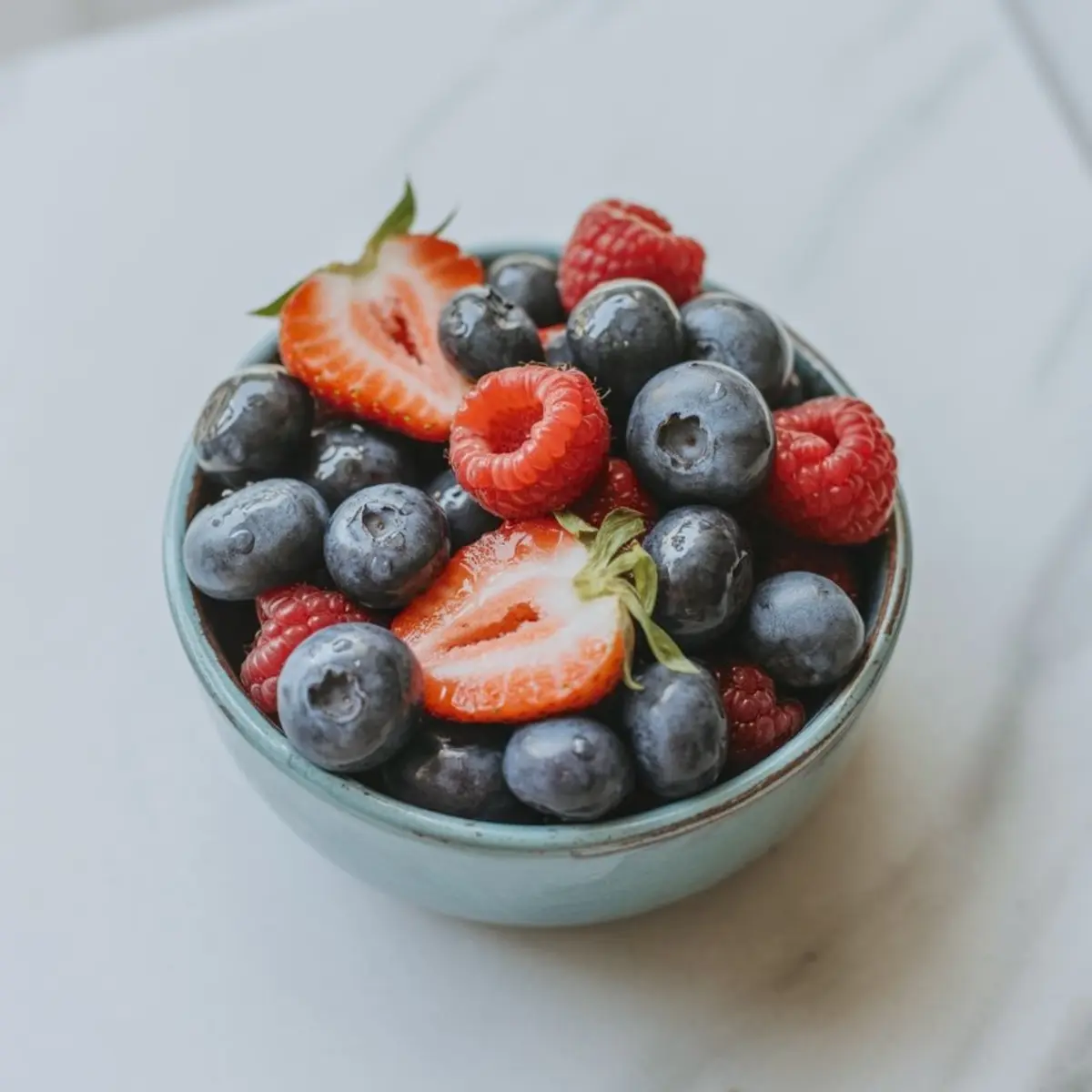 Close-up of a blue ceramic bowl filled with fresh blueberries, raspberries, and halved strawberries, placed on a white marble surface.