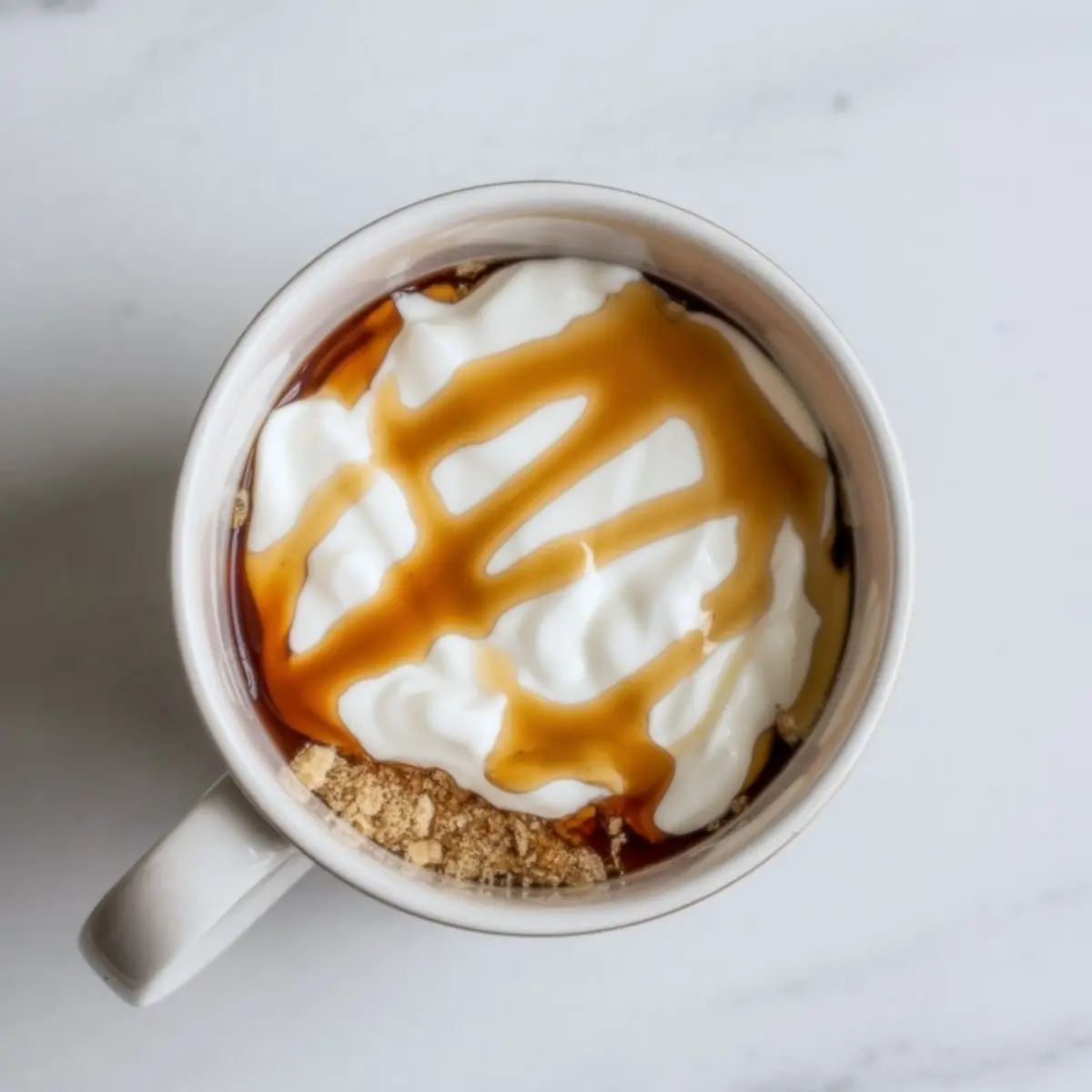 Top-down view of a mug cake topped with whipped cream, drizzled with caramel sauce, and sprinkled with crushed biscuits in a white ceramic mug on a light background.