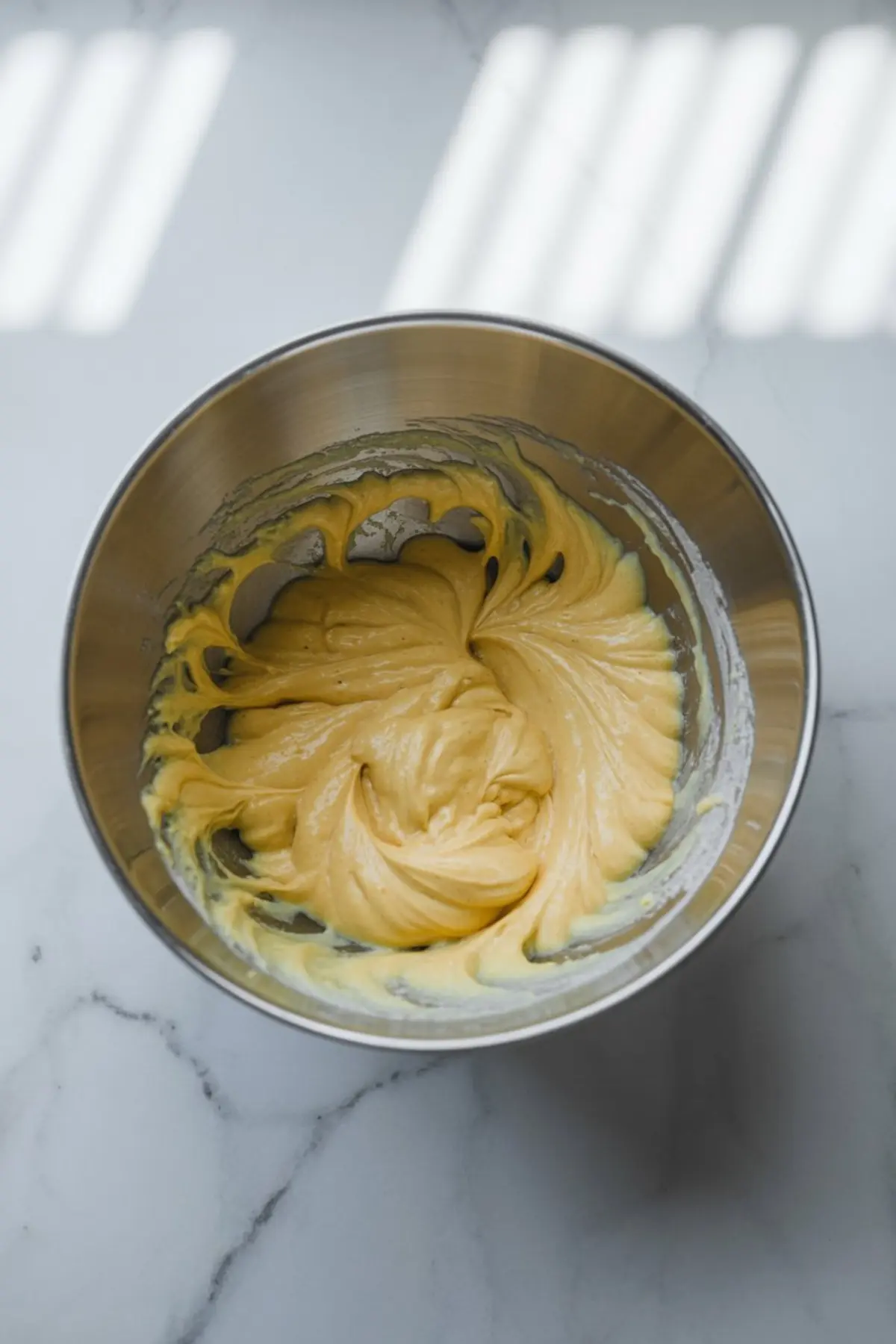 Yellow vanilla cake batter mixed to a smooth texture in a stainless steel bowl on a marble surface, ready for pouring into a baking pan.