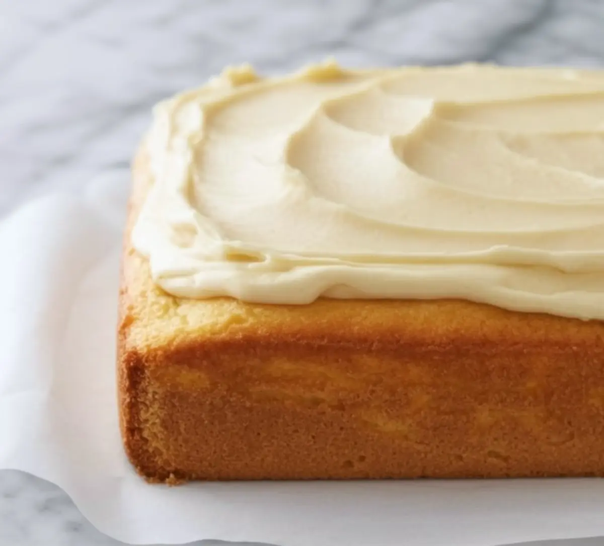 Freshly baked vanilla sheet cake topped with swirled vanilla frosting, shown from a side angle on a parchment-lined marble surface.