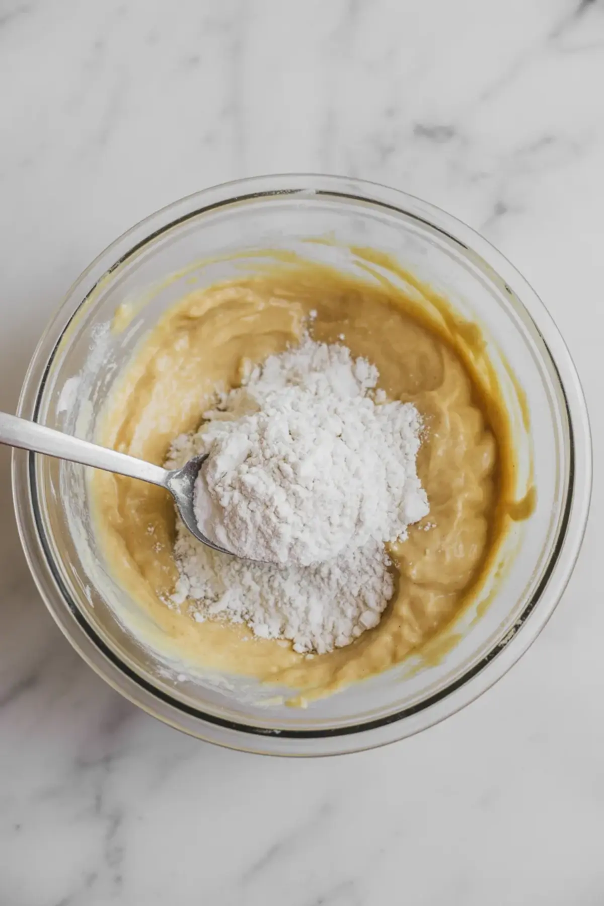 A spoonful of powdered sugar is held over a bowl of thick almond paste mixture, showing an in-process step of preparing almond croissant filling on a white marble surface.