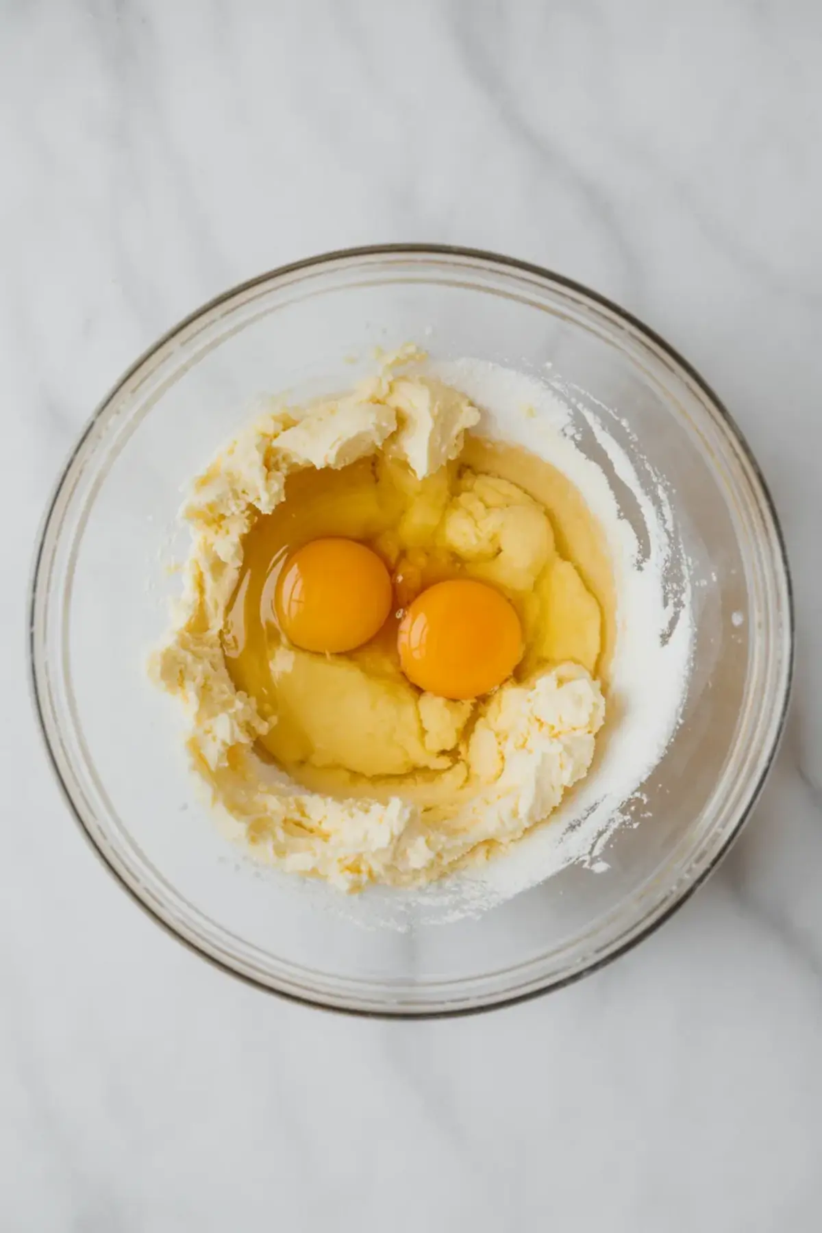 Two cracked eggs sit atop a partially mixed blend of butter and sugar in a glass bowl, capturing the egg addition step in making almond croissant filling.