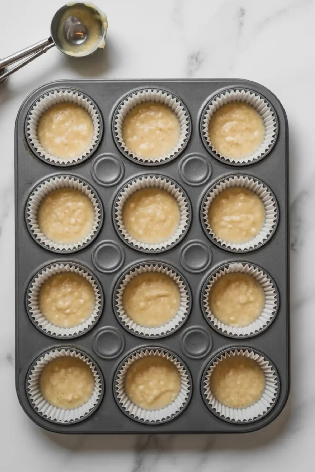 Muffin tin filled with raw almond flour batter in white paper liners, with a cookie scoop resting nearby on a white marble surface.