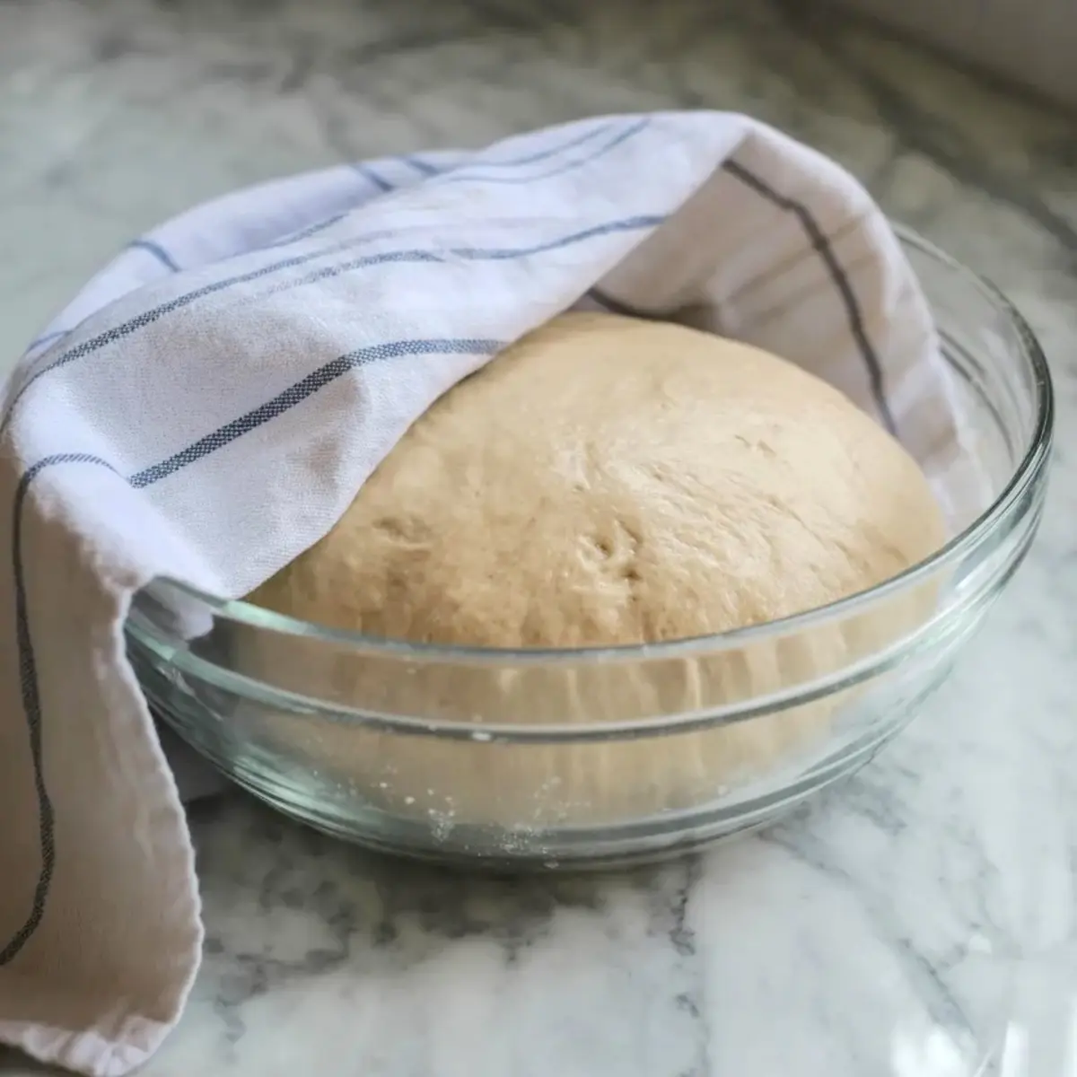 Soft risen dough rests in a glass bowl covered with a kitchen towel, showing smooth elastic texture for Italian bomboloni dough.