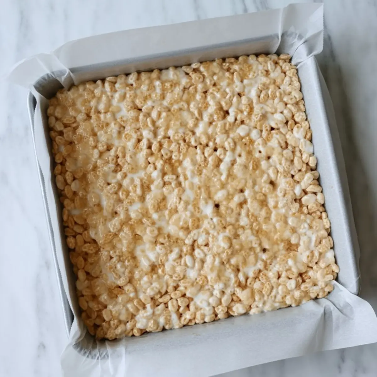 A square pan filled with freshly made Rice Krispies treats, featuring a glossy top layer of melted marshmallows and puffed rice cereal, set on a marble surface and lined with parchment paper.