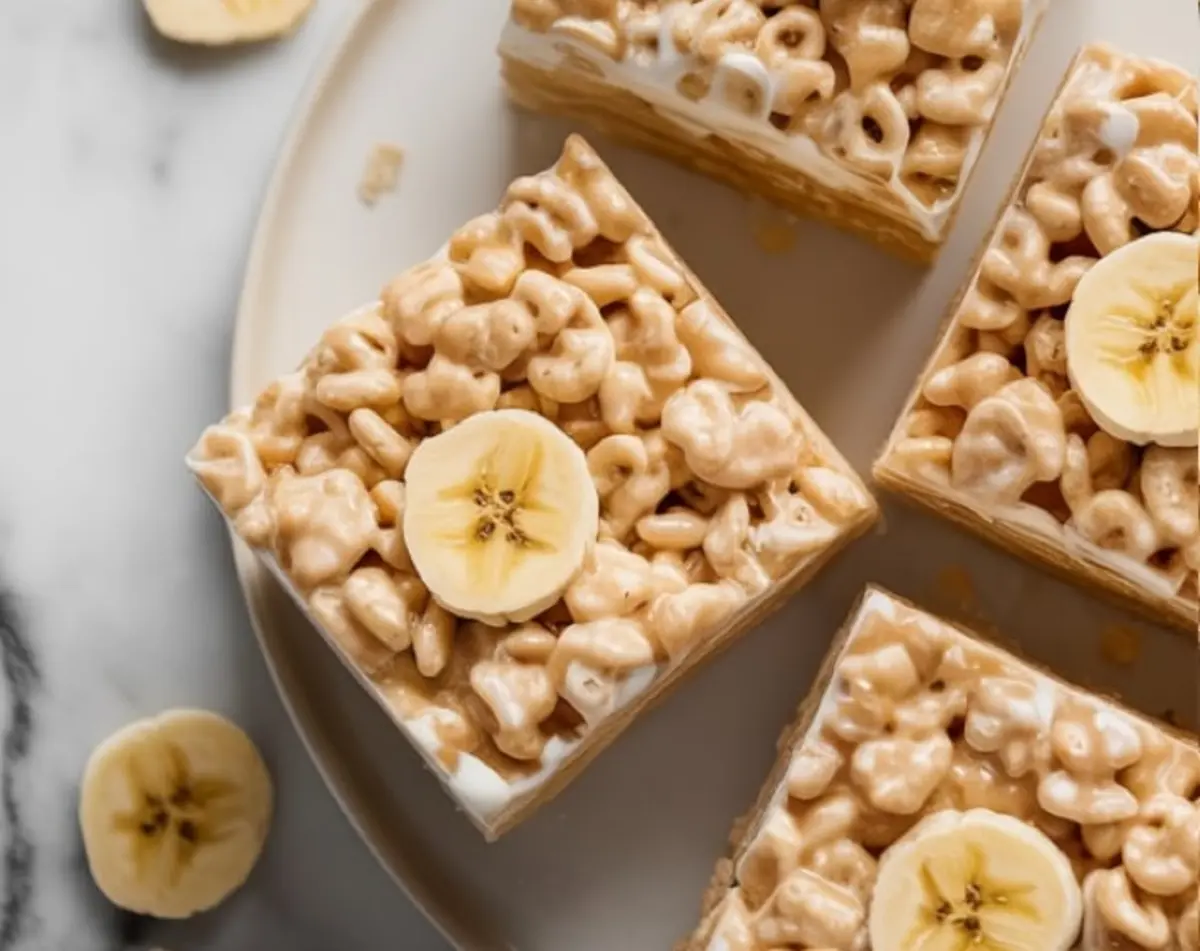 A top-down view of banana pudding Rice Krispies squares arranged on a round plate, each treat topped with a fresh banana slice and surrounded by additional banana rounds on a marble surface.