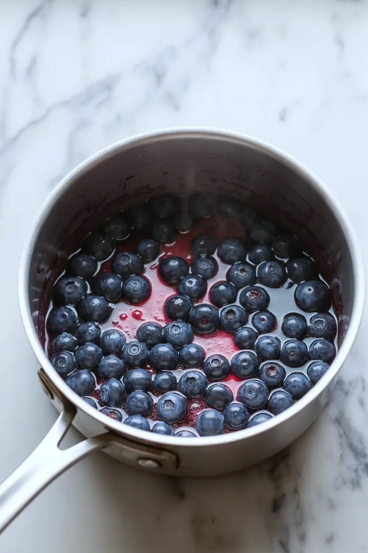 Overhead view of a stainless steel saucepan filled with fresh blueberries simmering in red liquid on a marble countertop.
