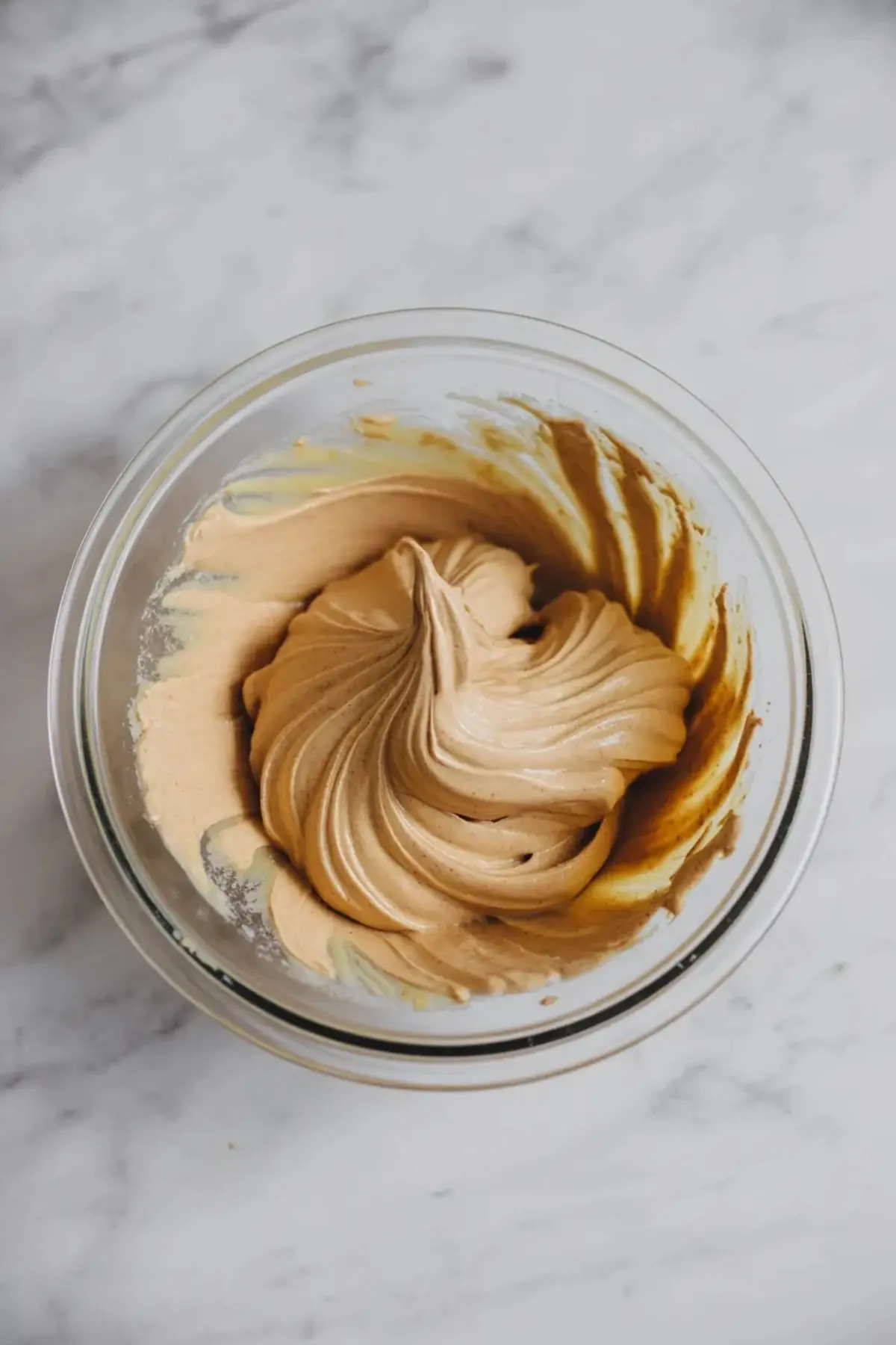 Fluffy whipped brown butter frosting with rich caramel tones in a glass mixing bowl, featuring a soft swirl pattern against a gray marble countertop.