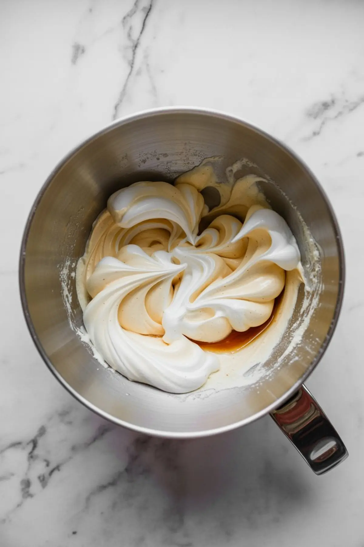 Fluffy egg whites folded into cookie batter in a silver mixing bowl, creating a swirled texture on a marble background.
