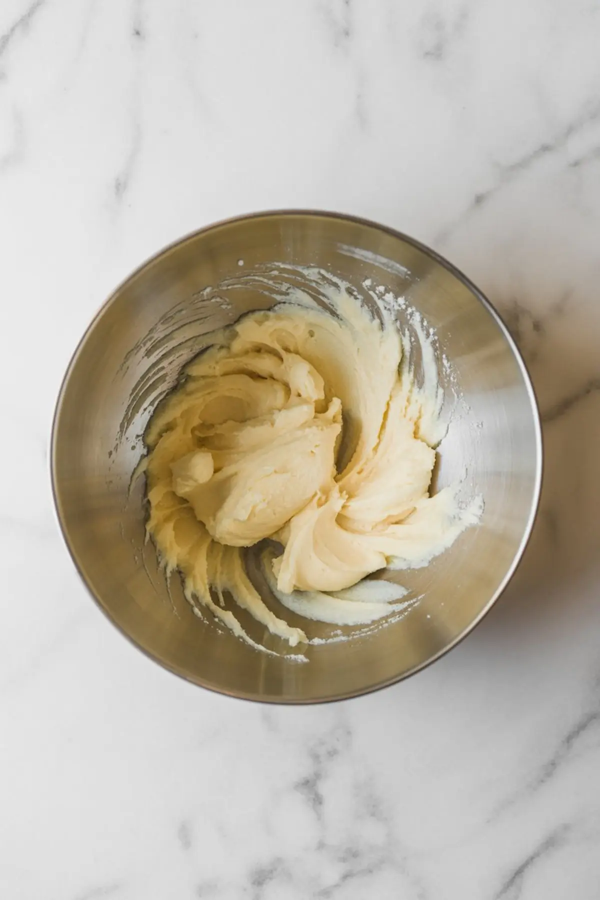 A stainless steel mixing bowl filled with creamy cookie dough on a white marble surface.
