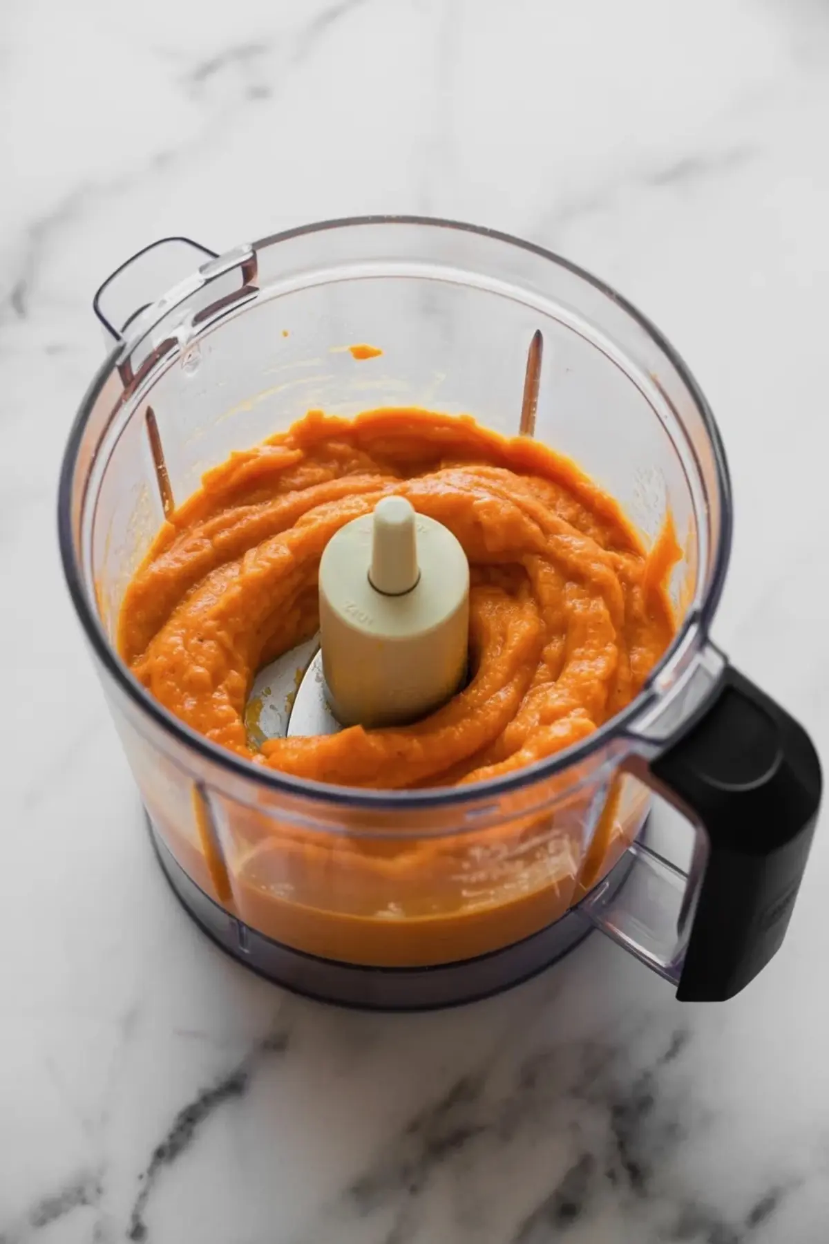 Smooth carrot puree inside a food processor bowl with white center blade, resting on a white marble countertop.