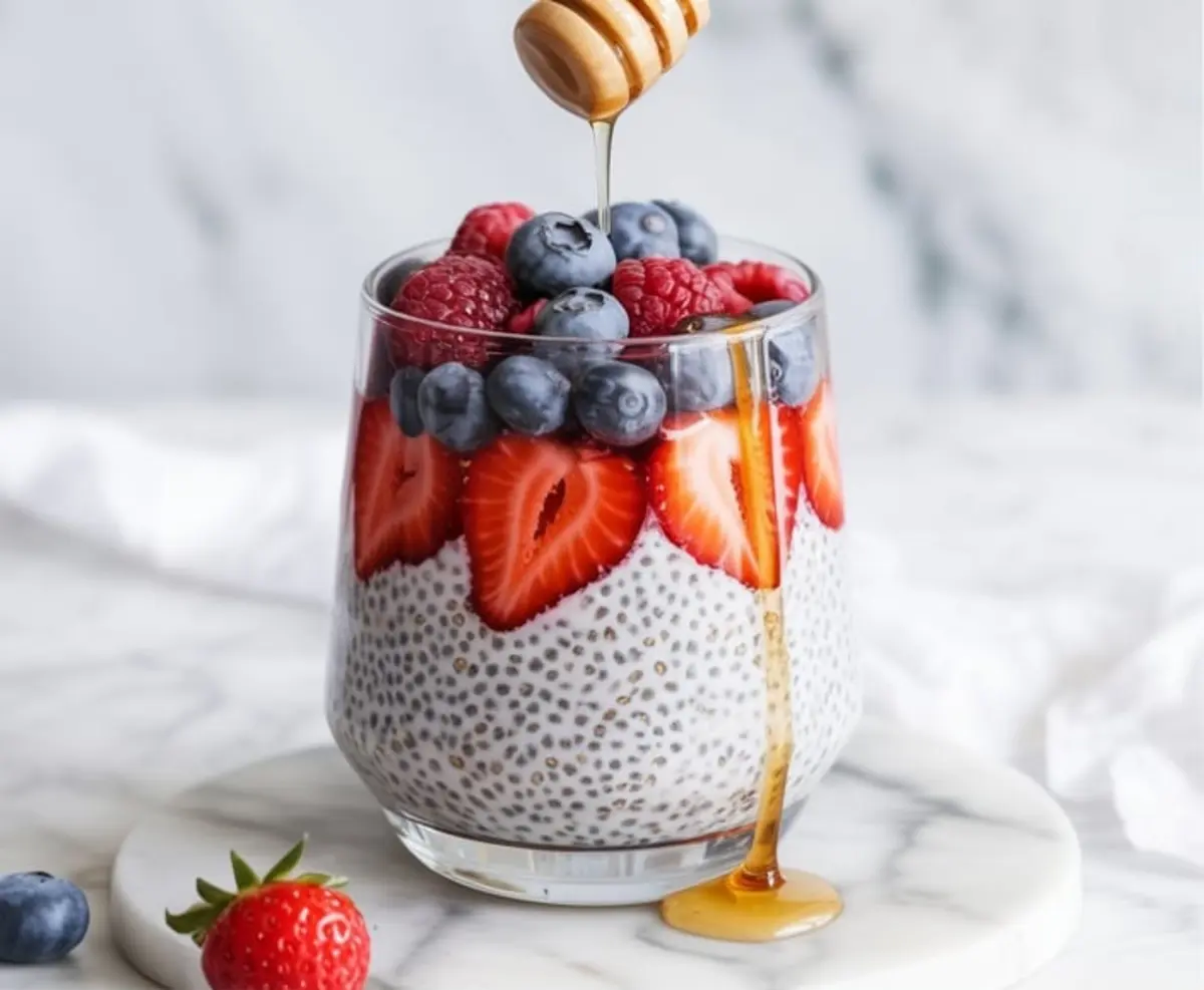 Side view of a glass filled with creamy chia pudding, layered with sliced strawberries, blueberries, and raspberries. Honey is being drizzled over the berries, flowing down the side of the glass onto a round marble board.
