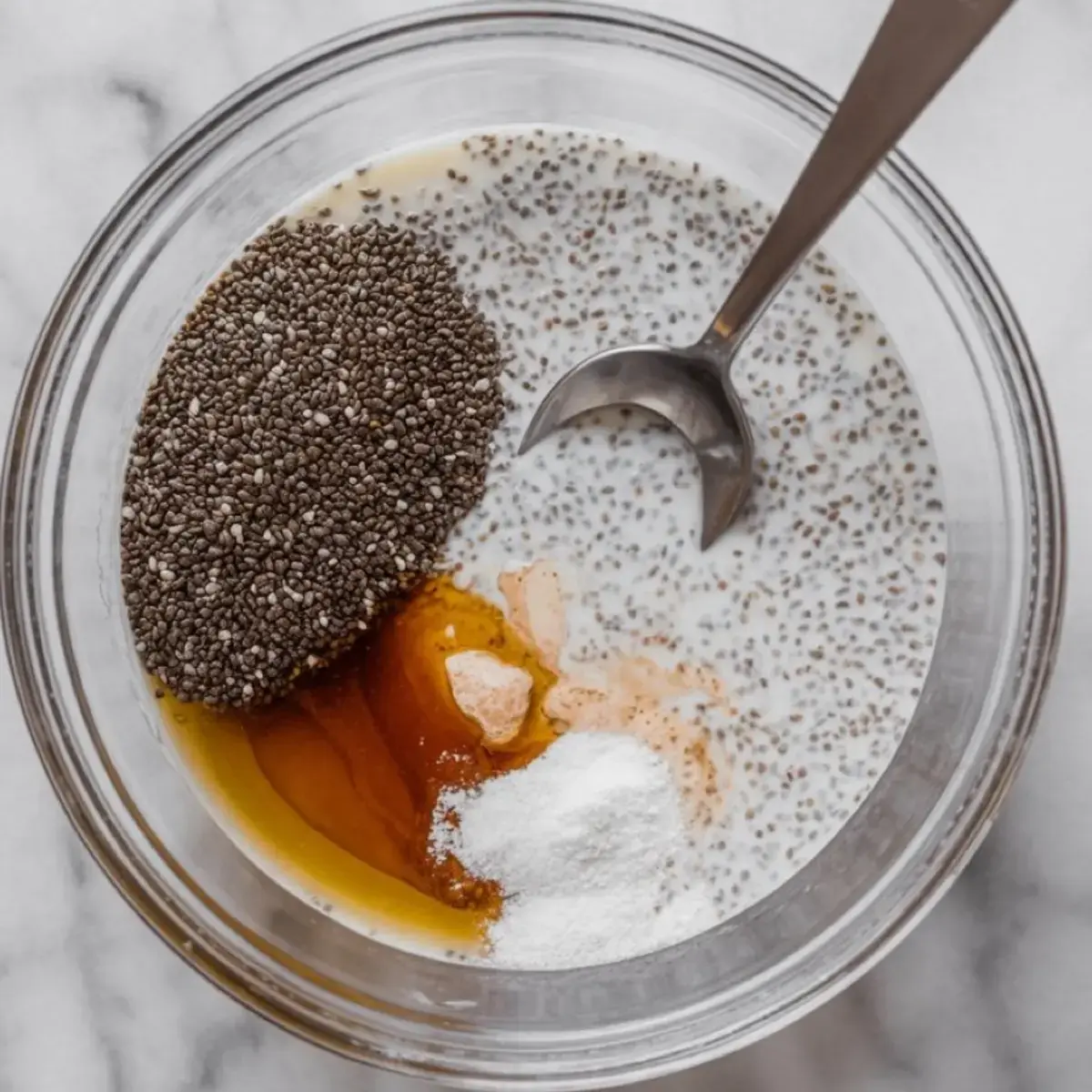 Close-up of a mixing bowl containing the ingredients for chia pudding, including chia seeds, honey, vanilla extract, sweetener, and milk. A metal spoon rests on the side, ready to combine the ingredients into a creamy mixture.
