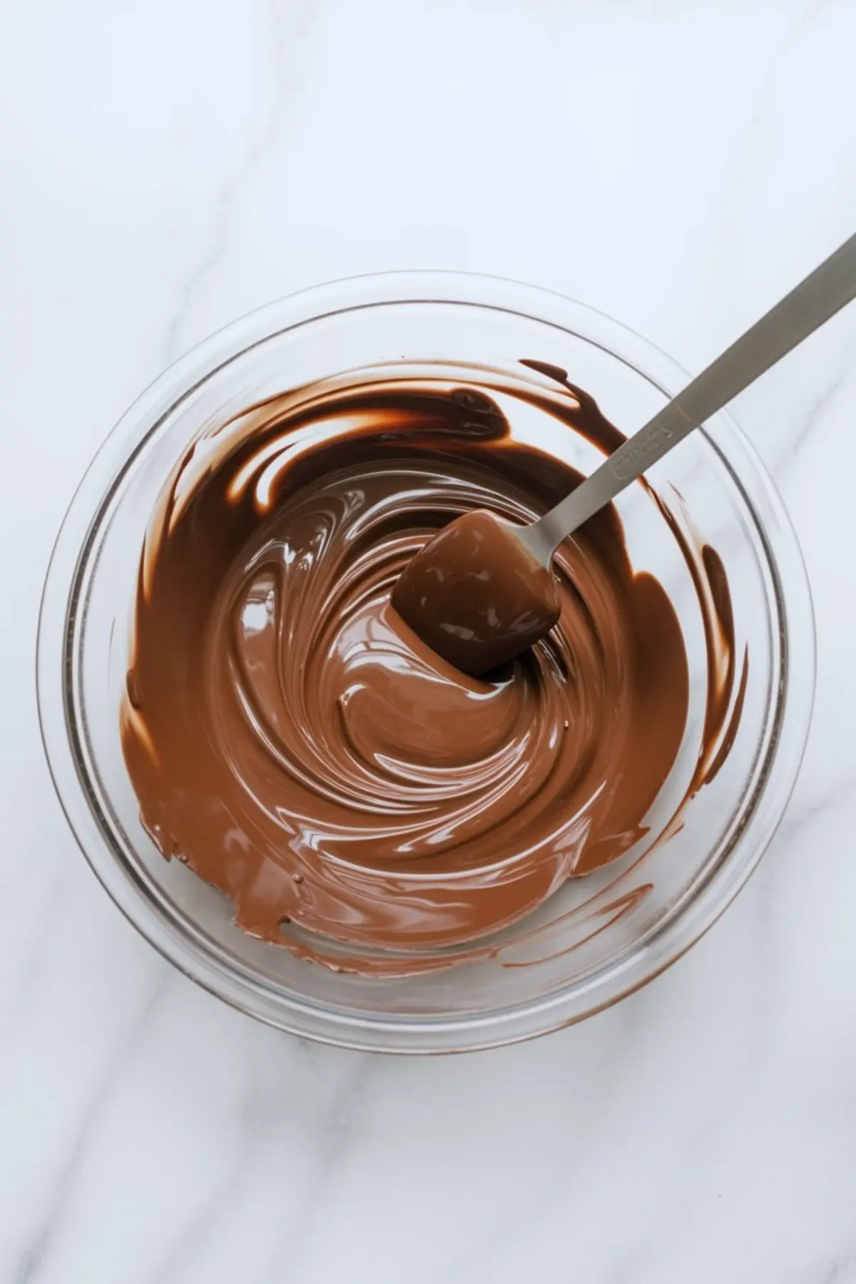 Swirled milk chocolate ganache in a glass bowl with a spatula, set on a white marble counter. Smooth melted chocolate texture ideal for cake fillings or dessert toppings.