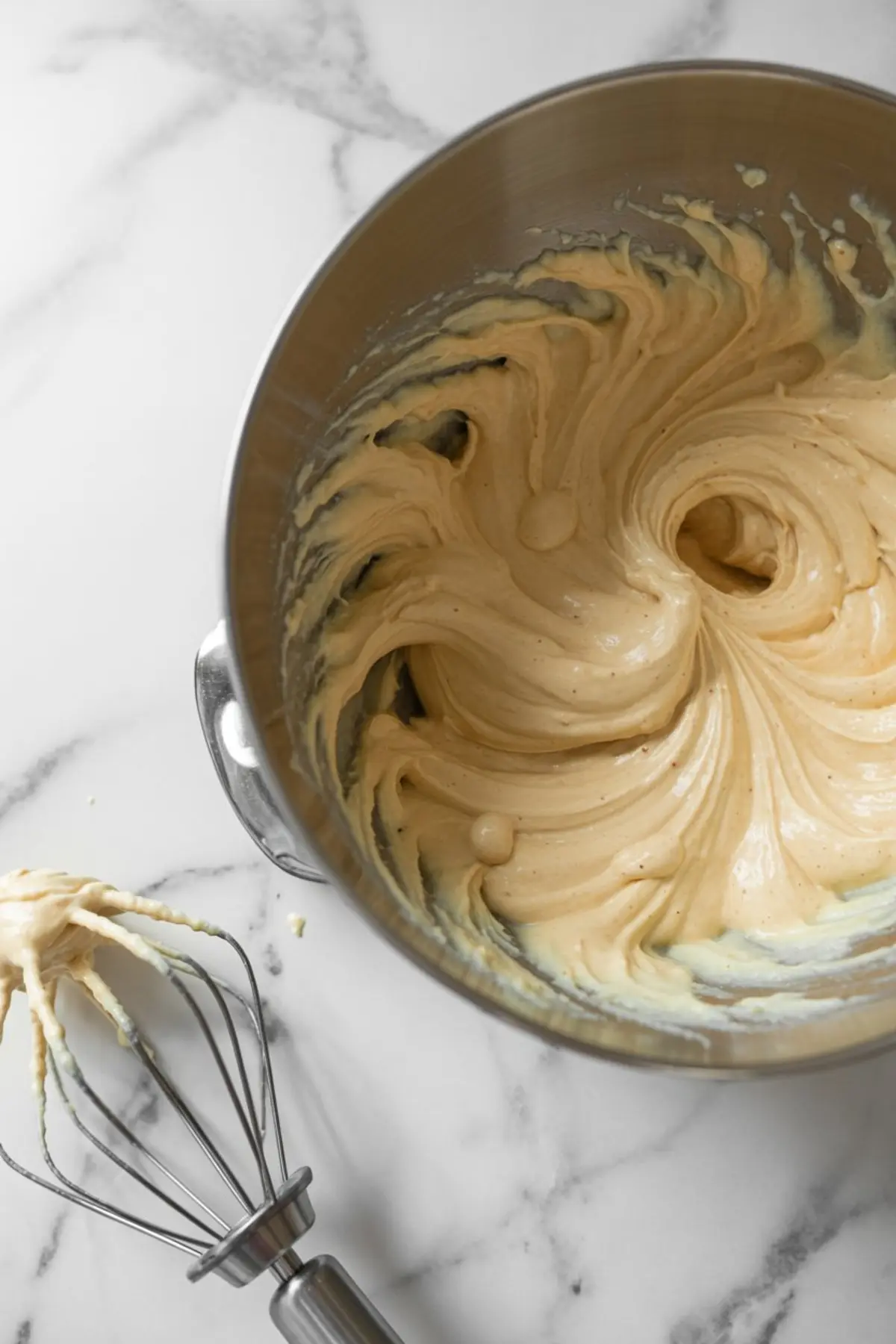 Overhead view of thick cheesecake batter whipped in a metal mixing bowl with a whisk attachment resting beside the bowl on a marble surface.