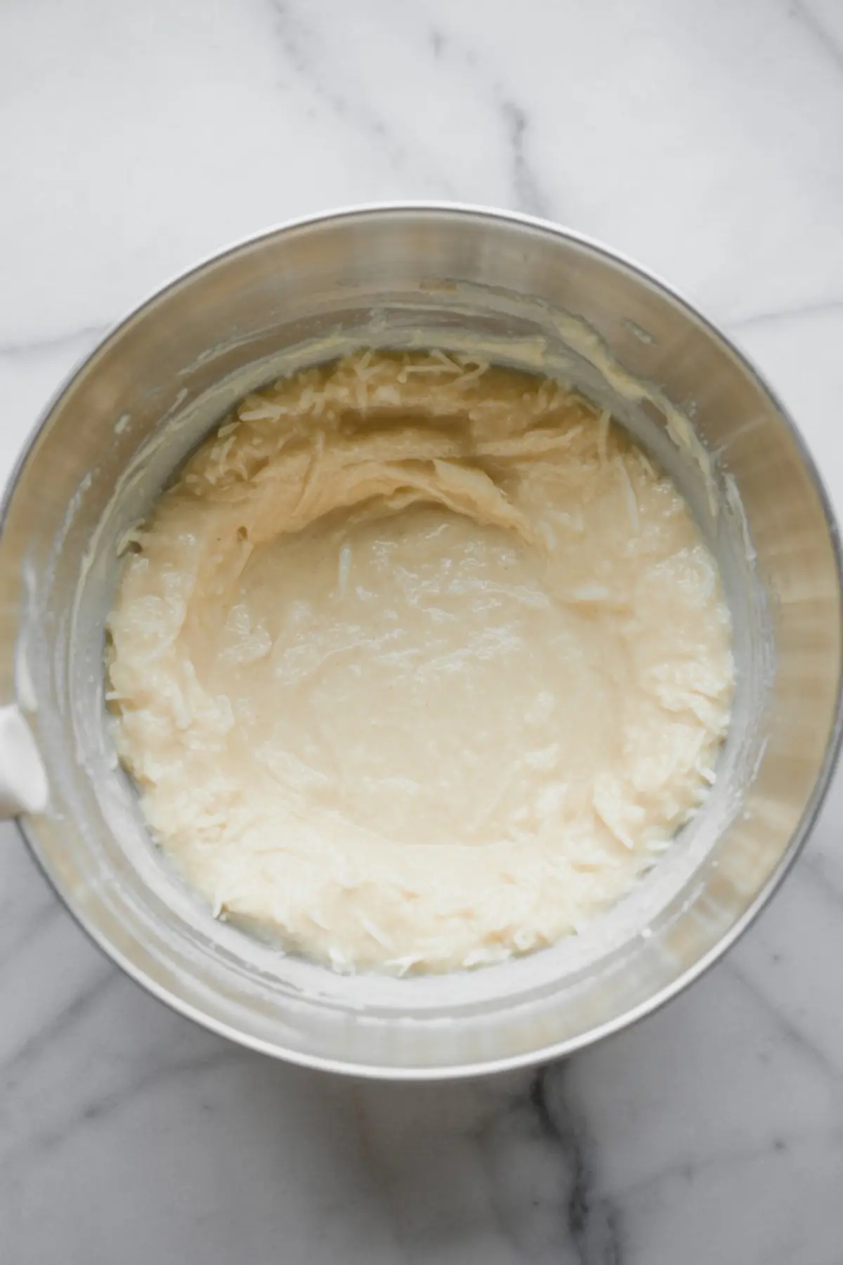 A stainless steel mixing bowl contains a thick coconut filling mixture with visible shredded coconut, set against a white marble background.
