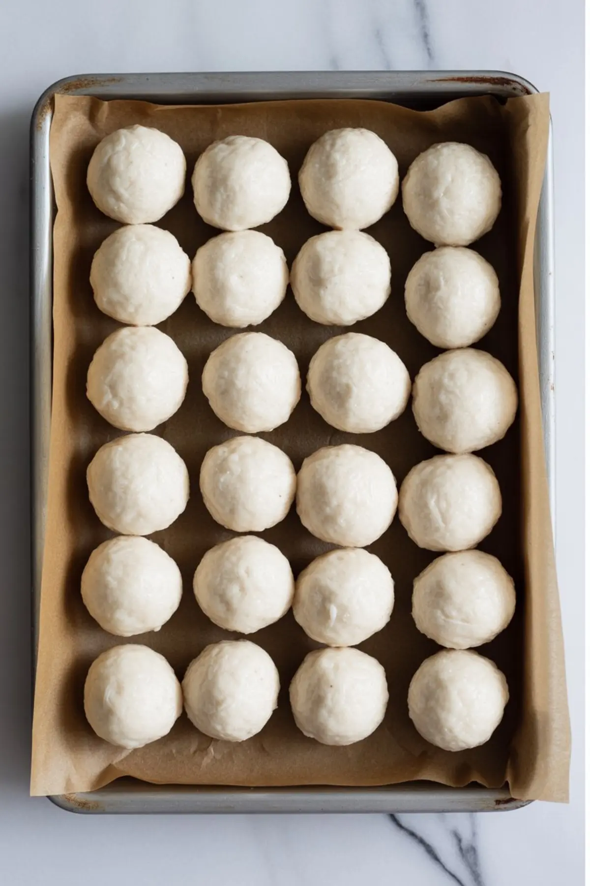 A parchment-lined baking sheet holds neatly arranged unbaked dough balls in a 5x5 grid, ready for baking on a clean marble countertop.
