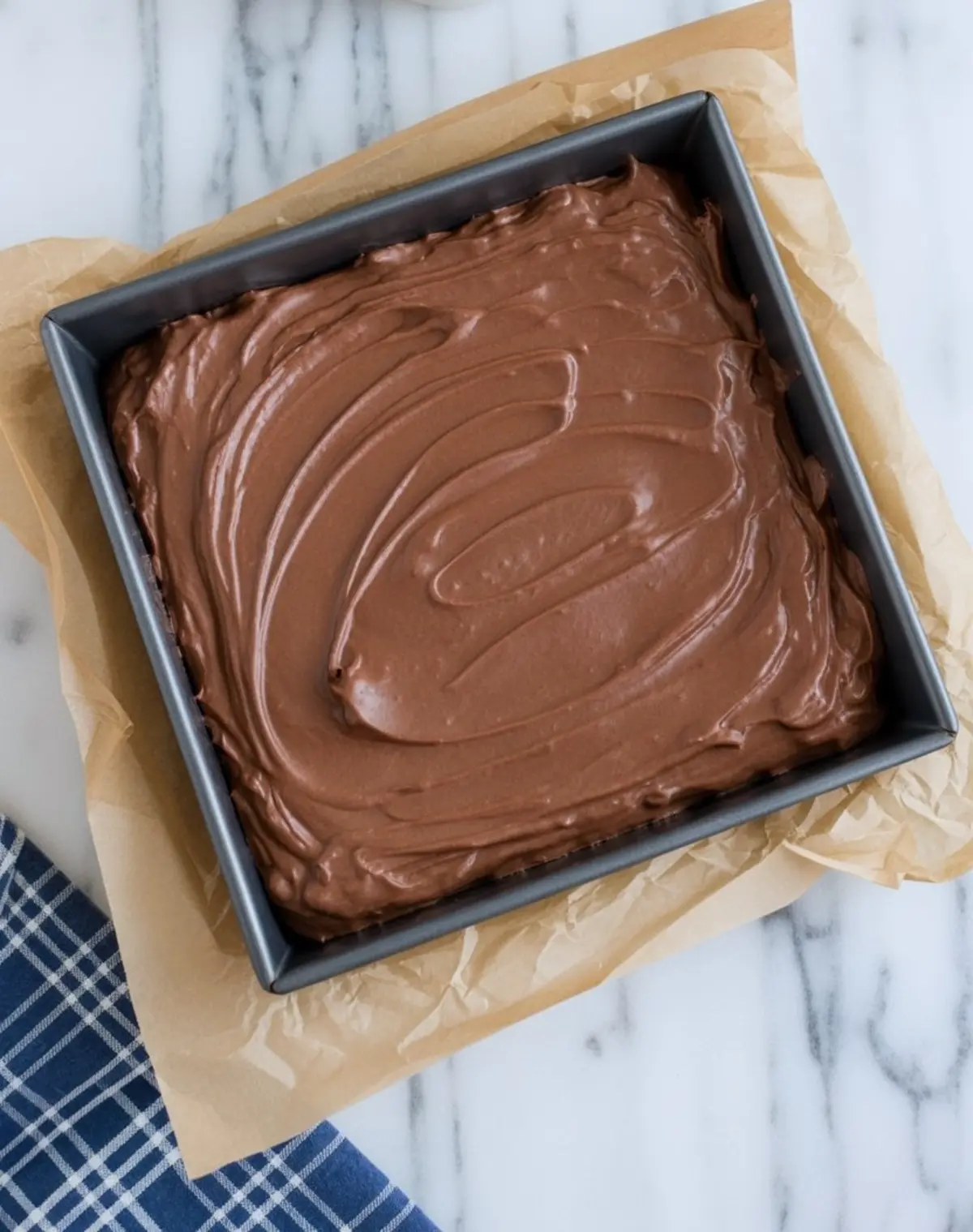 Chocolate fudge mixture spreads smoothly inside a square baking pan lined with parchment paper on a marble countertop.