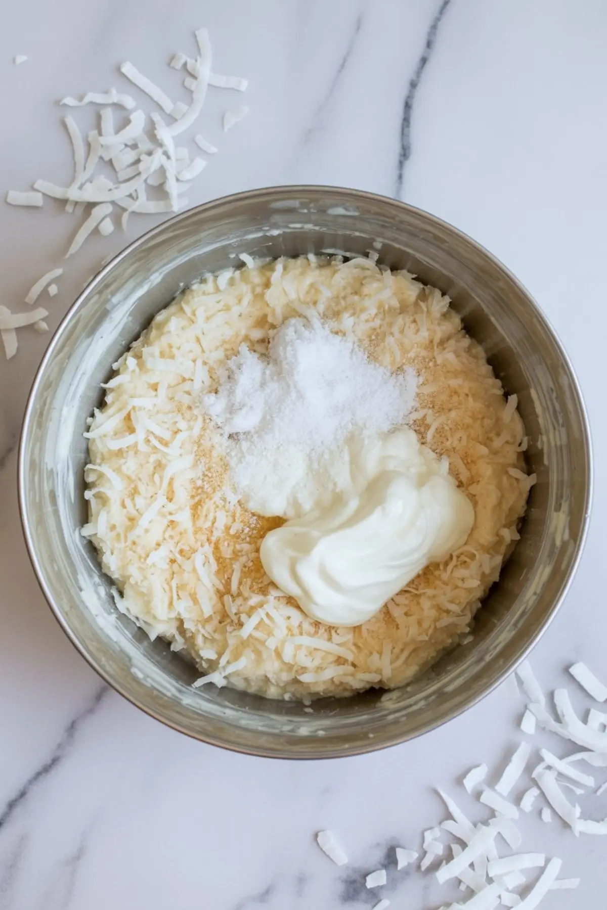 Metal mixing bowl filled with shredded coconut, sour cream, sugar, and vanilla, surrounded by scattered coconut flakes on a marble surface.