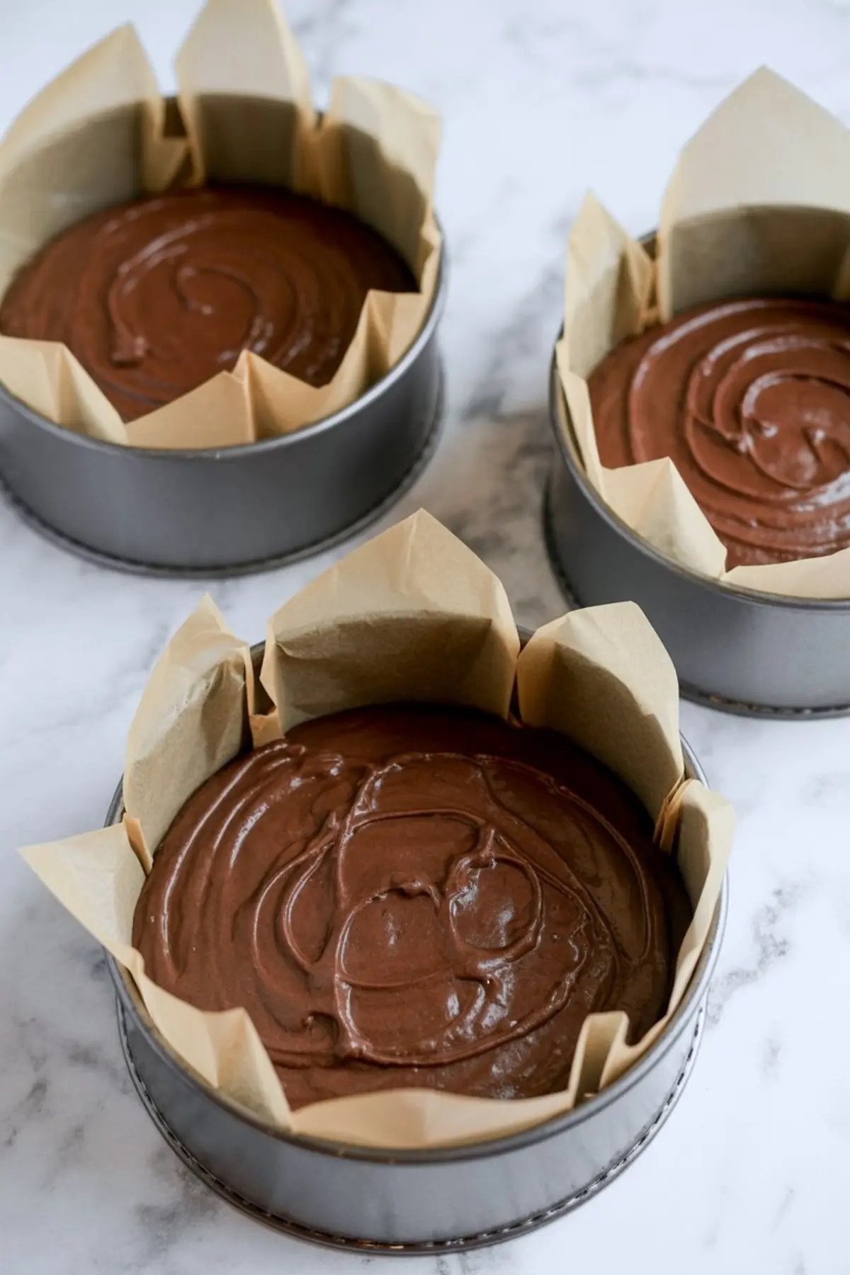 Three round baking pans lined with parchment paper, each filled with rich chocolate cake batter ready to bake.