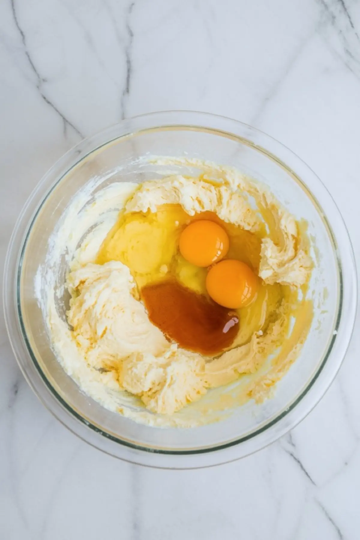A clear glass bowl with creamed butter, sugar, vanilla extract, and two raw eggs, set on a white marble countertop.
