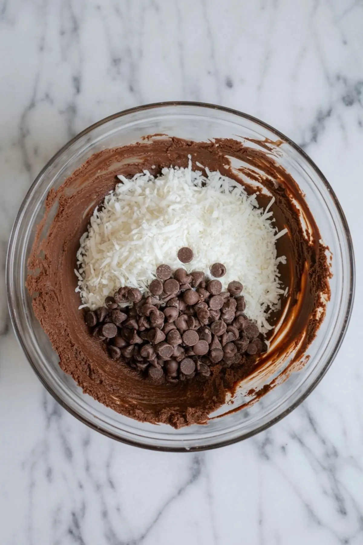 Glass mixing bowl filled with chocolate cookie dough topped with shredded coconut and chocolate chips on a white marble counter.
